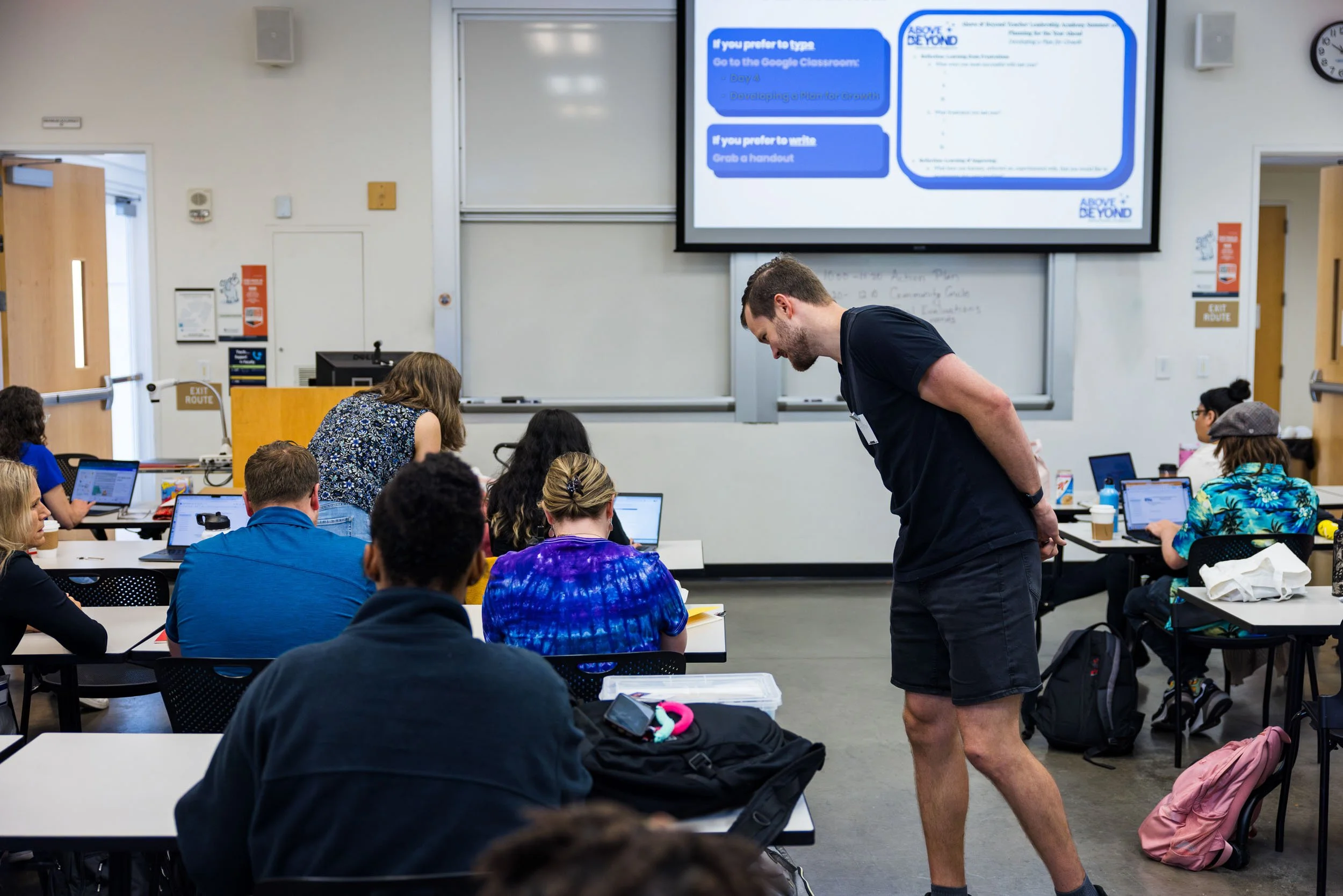 A classroom with students sitting at desks using laptops, a teacher assisting a student, and a large screen displaying a presentation on the wall.
