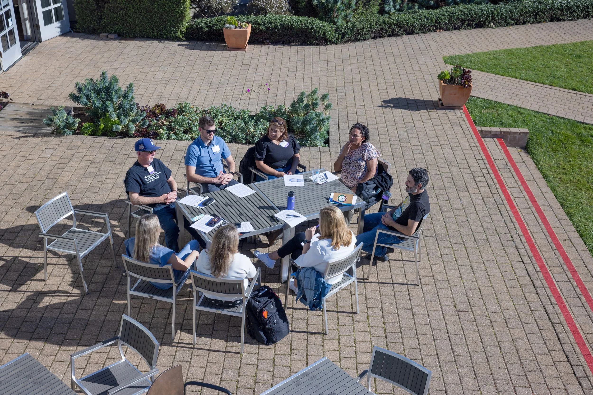 A group of nine people seated around a rectangular outdoor table with papers, notebooks, and drinks, engaging in a discussion on a brick-paved patio surrounded by plants and greenery.
