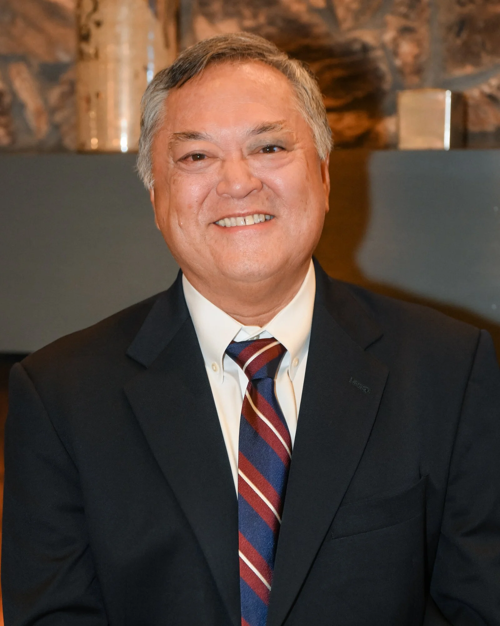 A smiling middle-aged man in a black suit with a striped tie, sitting indoors with a wooden background.