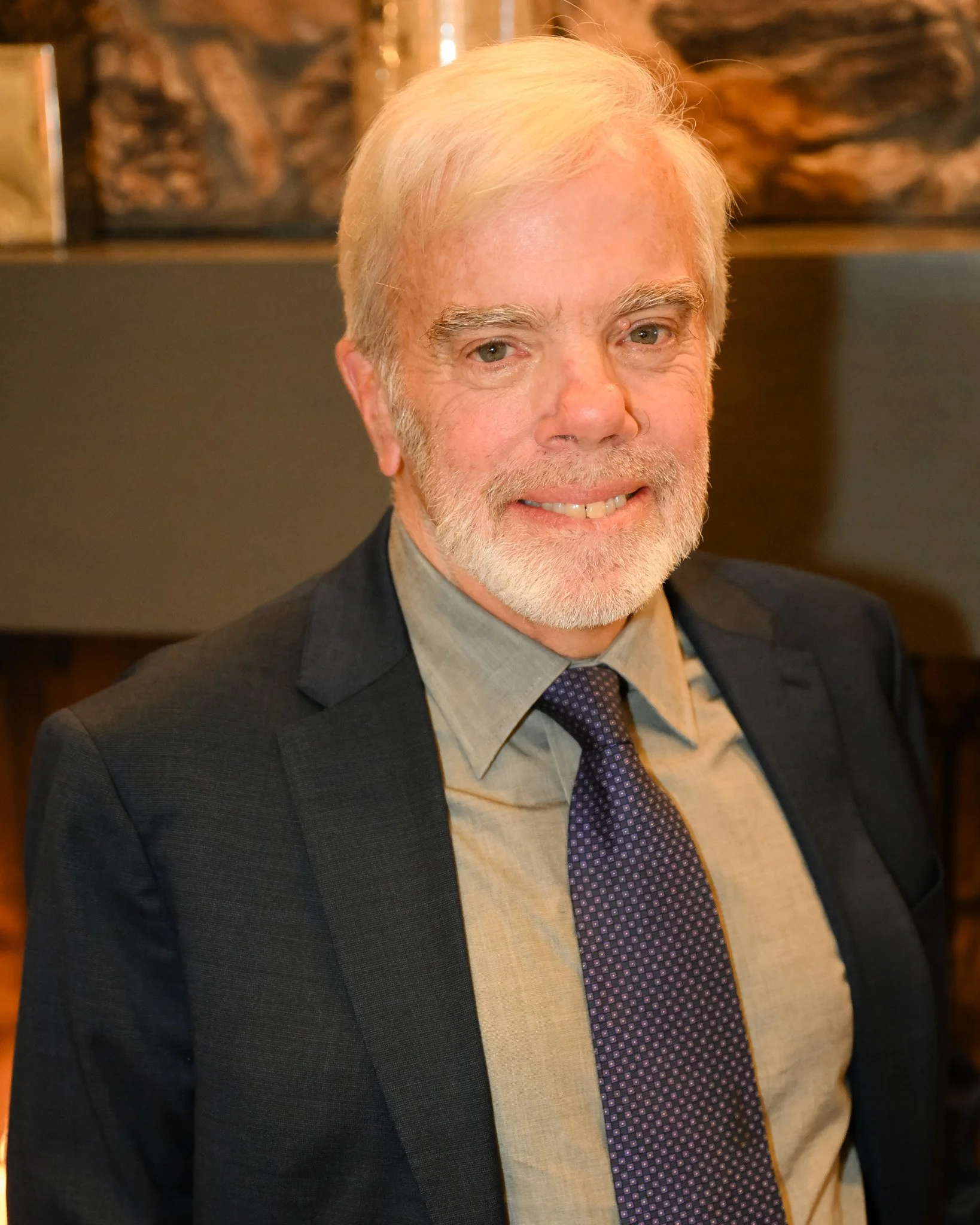 An elderly man with white hair and a beard smiling, dressed in a suit and tie, standing indoors with a blurred background.