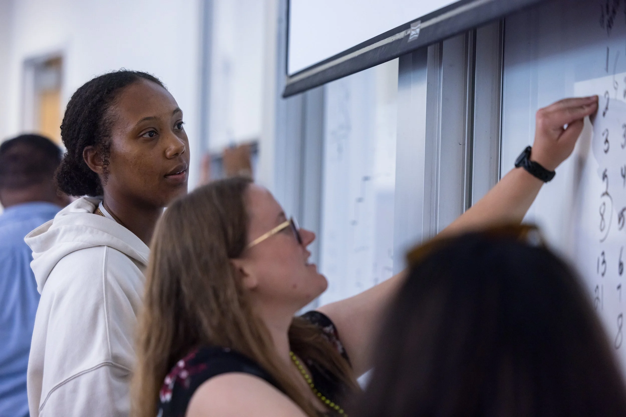 Two women standing near a whiteboard, one writing on it and the other looking on, in a classroom or seminar setting.