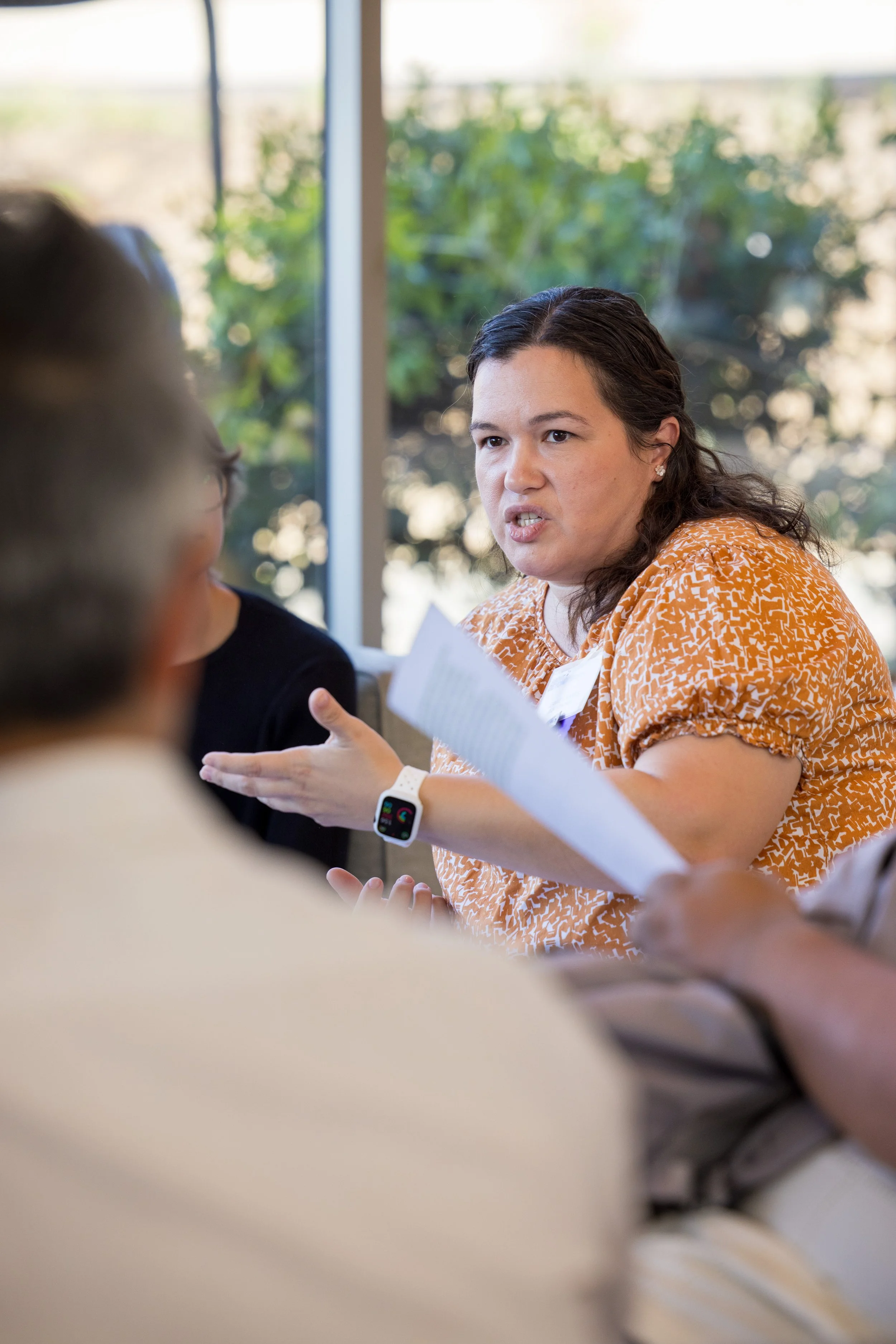 Woman in orange patterned blouse talking passionately during a discussion in a bright room with large windows and trees outside.