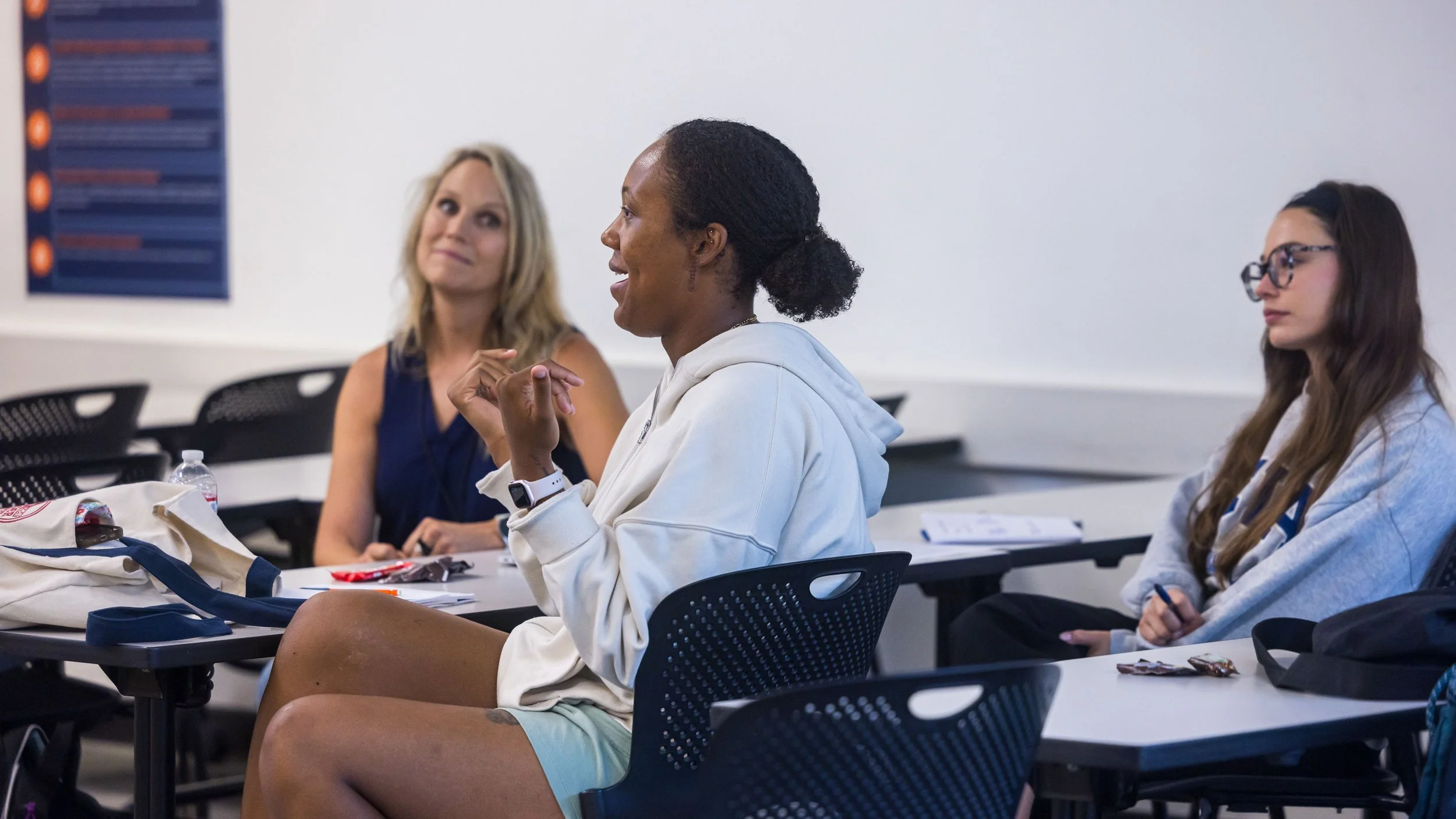A group of women sitting in a classroom, engaging in a discussion or listening to a speaker. The woman in the center is wearing a white hoodie and shorts, smiling and gesturing with her hands. The women on either side are listening attentively, one with glasses and the other with blonde hair. There are backpacks, papers, and a water bottle on the desks.
