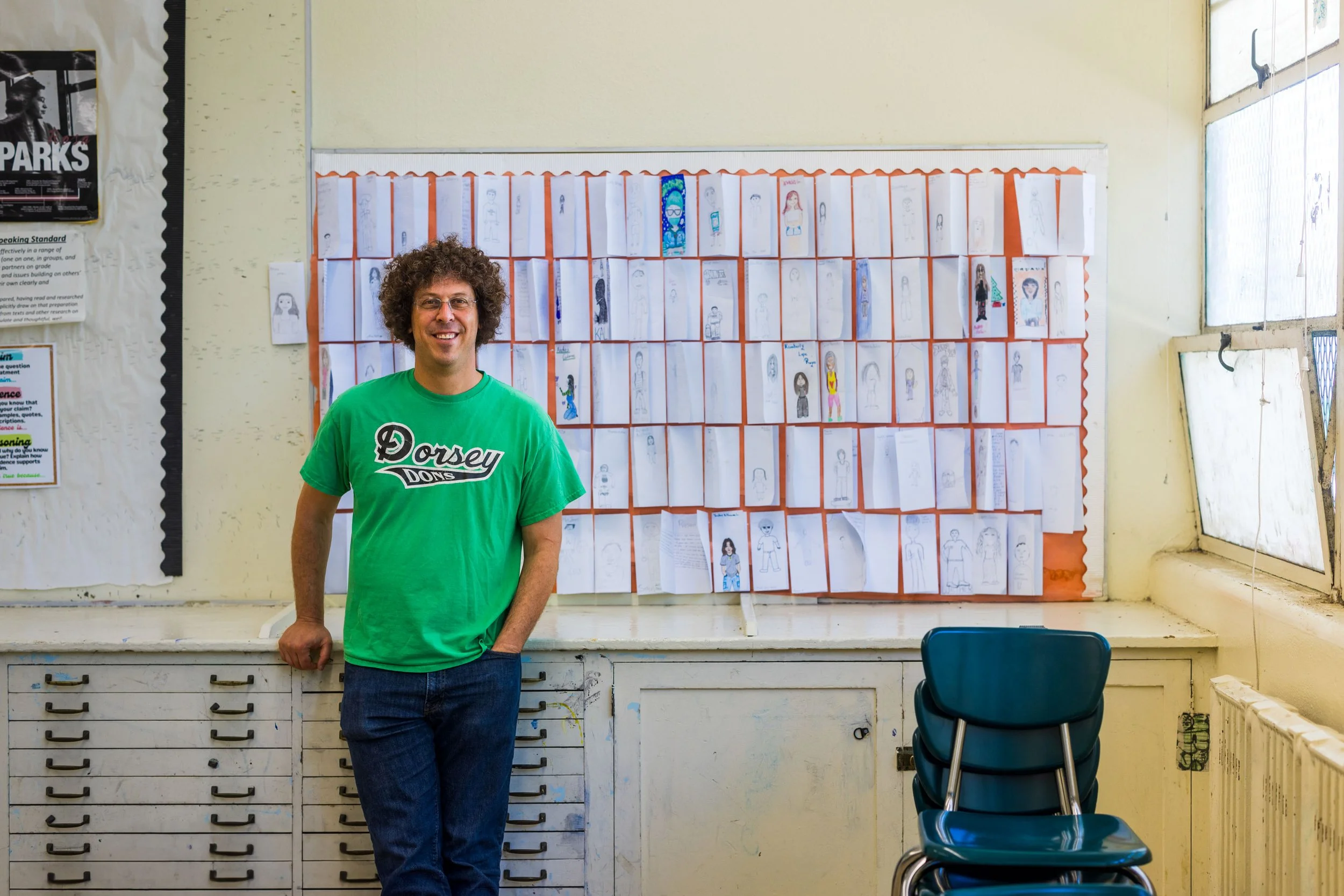 A man with curly hair wearing a green Dorsey Dons t-shirt, standing in front of a wall of children's drawings on paper held by orange borders inside a classroom.