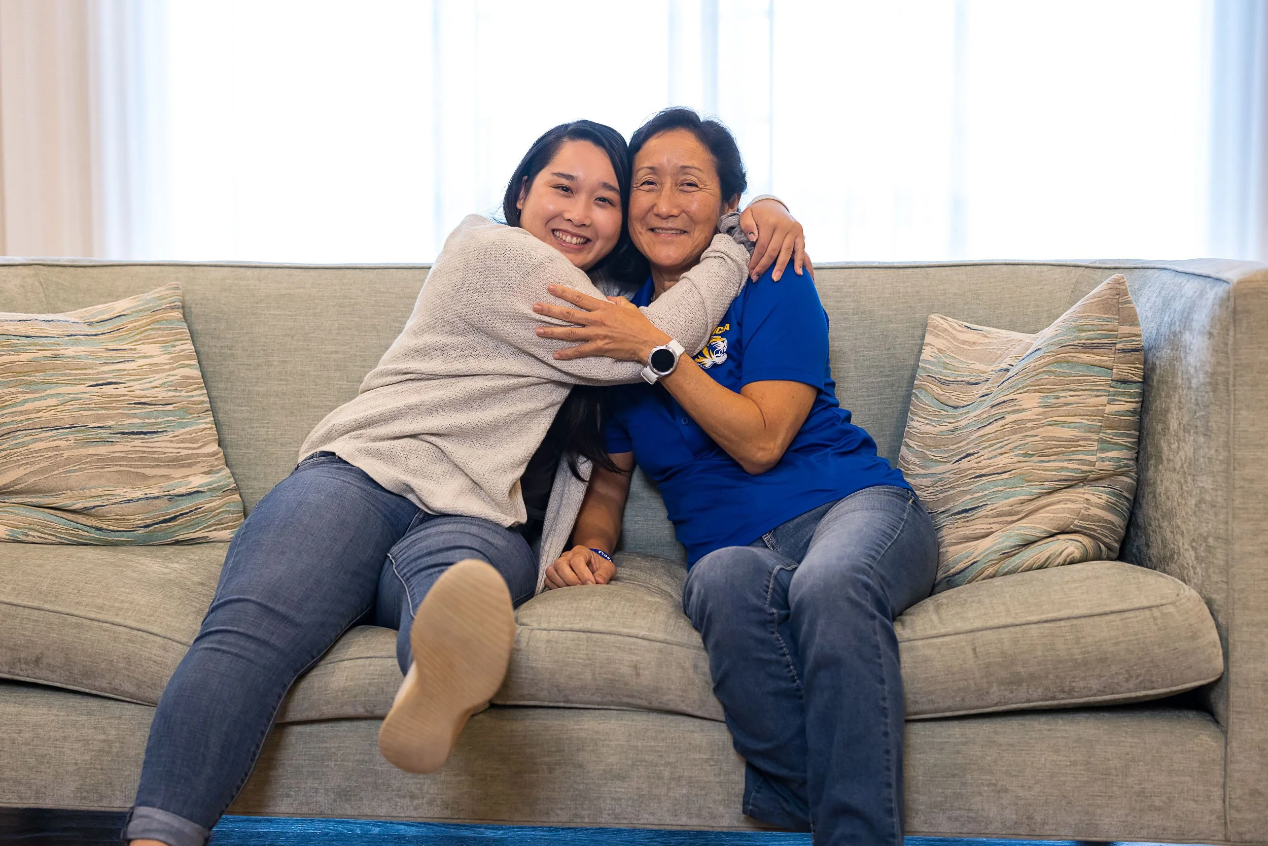 Two women sitting on a beige couch, embracing and smiling indoors with natural light coming through the window behind them.