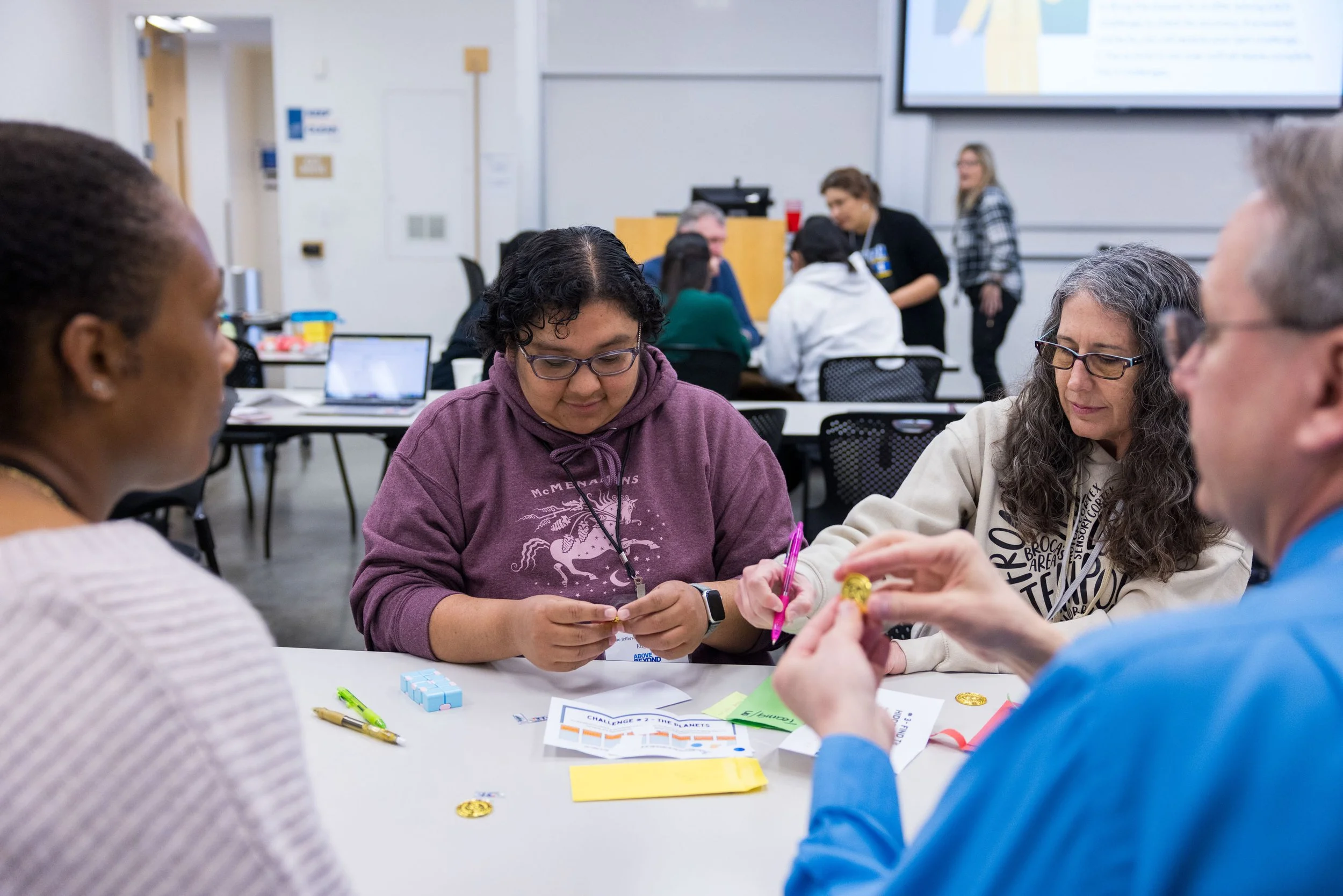 A group of four people sitting around a table in a classroom, engaging in an activity with papers, pens, and small objects, with others working in the background.