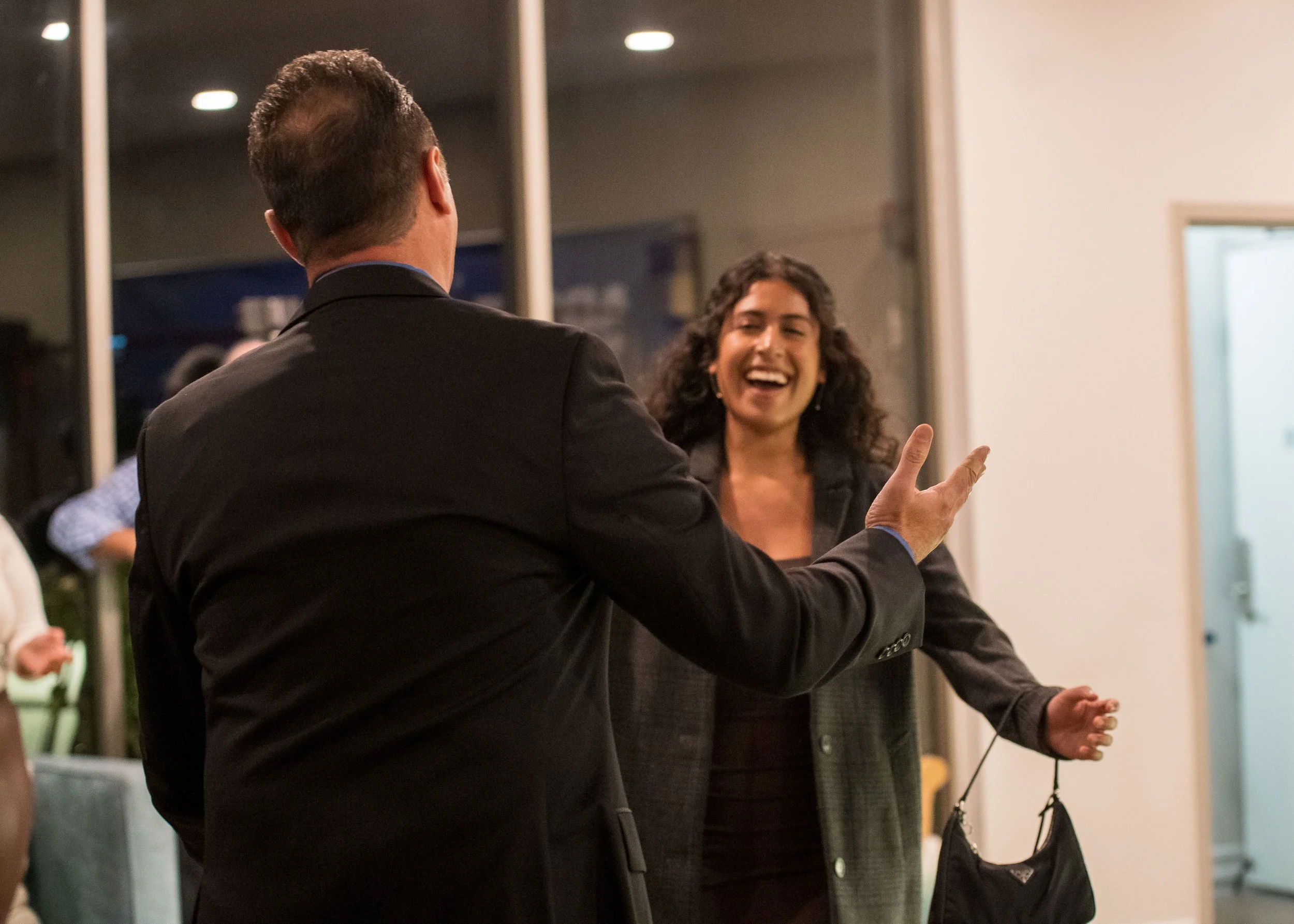 A man in a black suit is greeting a woman with curly hair, wearing a dark top and blazer, who is smiling and holding a handbag.