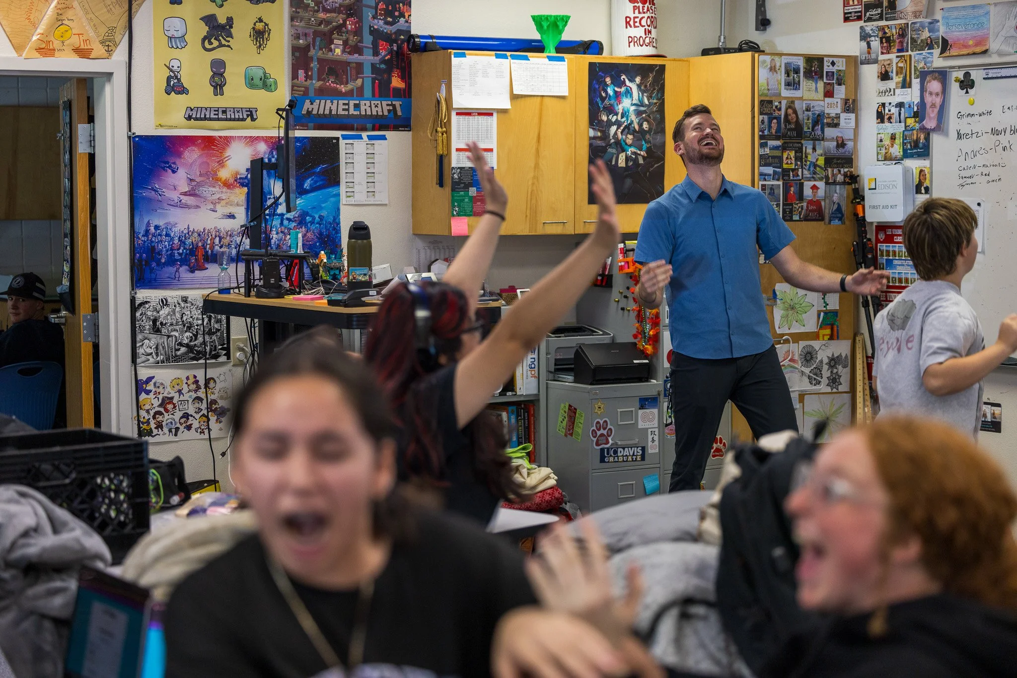 People in a classroom celebrating and having fun, with one person standing and others sitting or lying down, colorful posters and decorations on the walls.