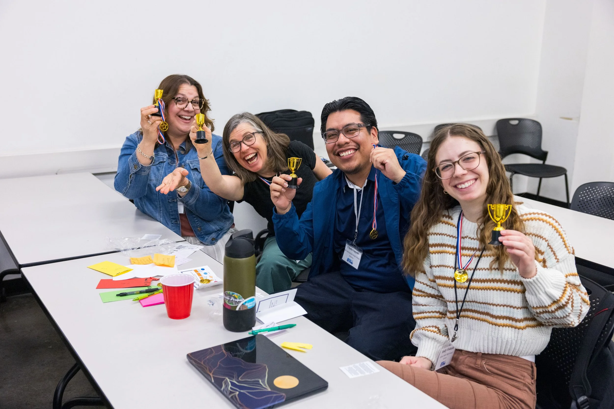 Five people sitting at a table celebrating, holding small gold trophies, wearing medals, and smiling with joy.