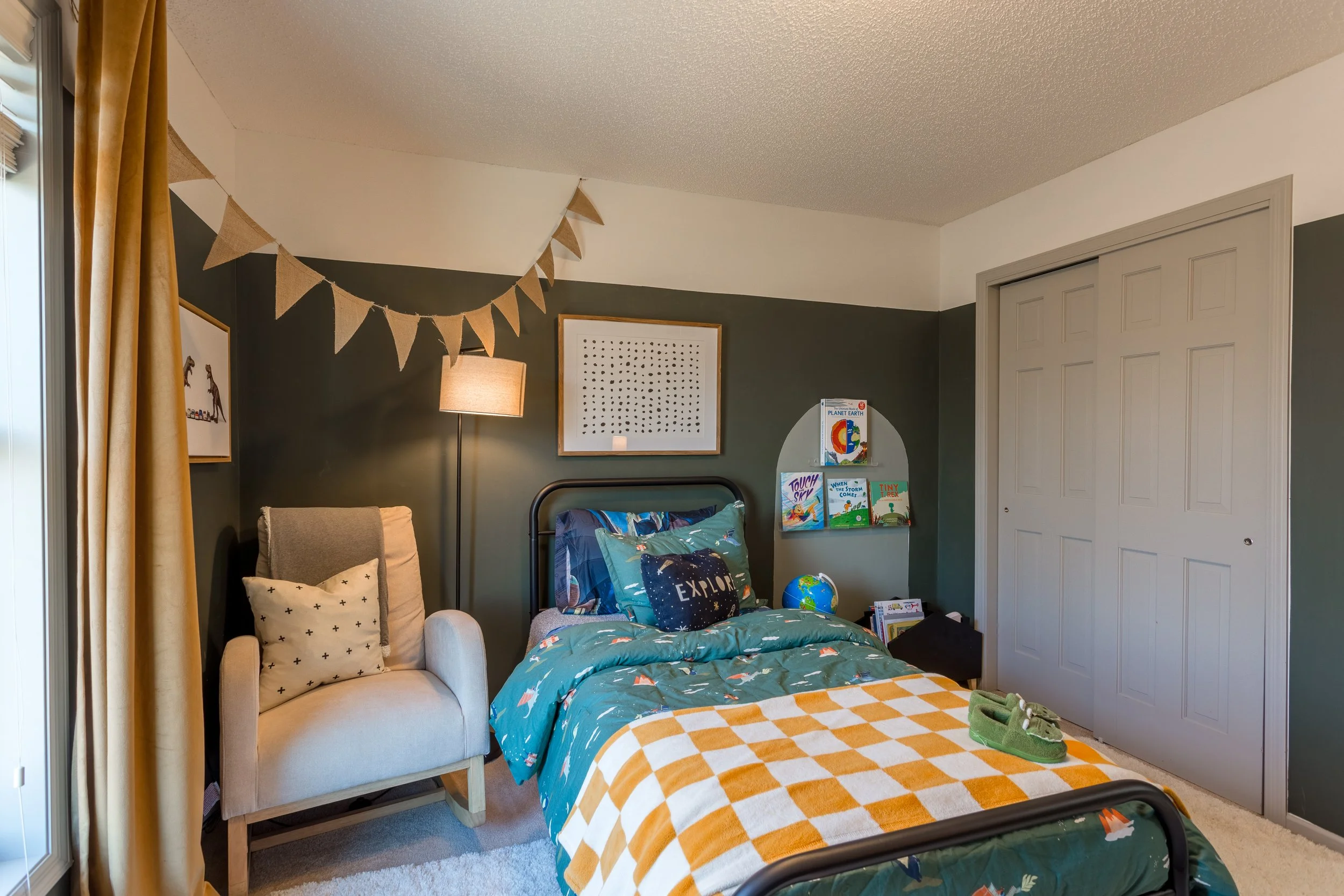 Child's bedroom with a bed featuring space-themed bedding, a chair with pillows, a lamp, wall art, books, a globe, and a pair of green shoes on the bed. Natural light coming through a window with gold curtains.