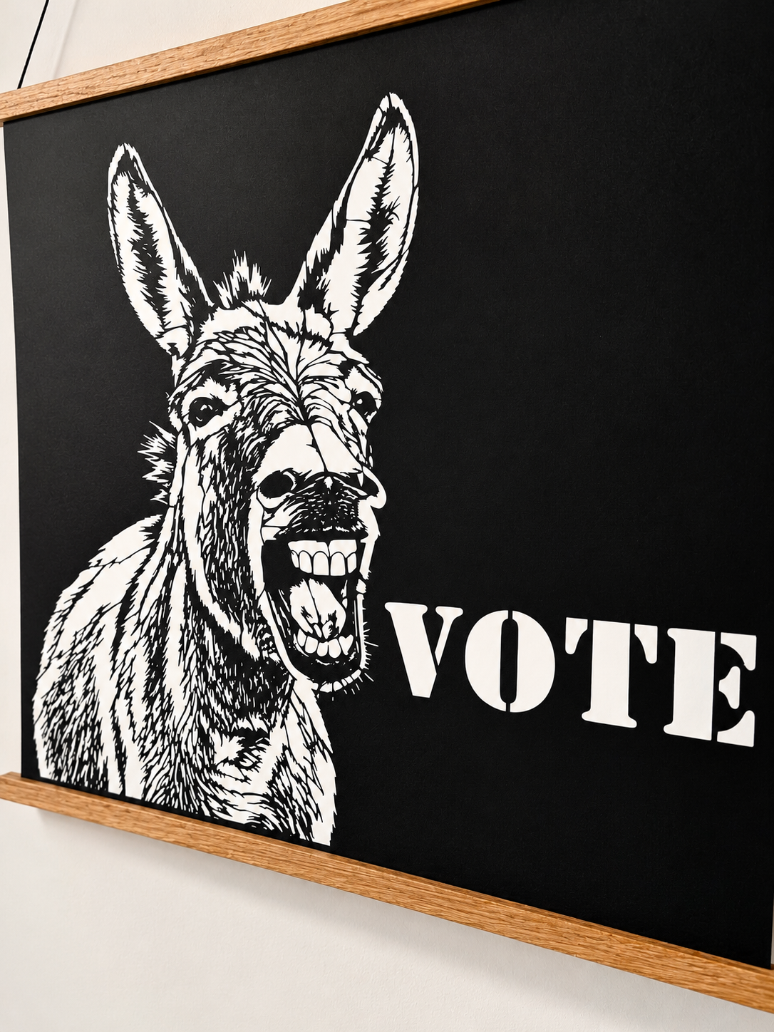 Writing       Ruthie political donkey VOTE poster, original black cut paper wall art hanging at an angle from a wooden poster hanger against a neutral studio wall.