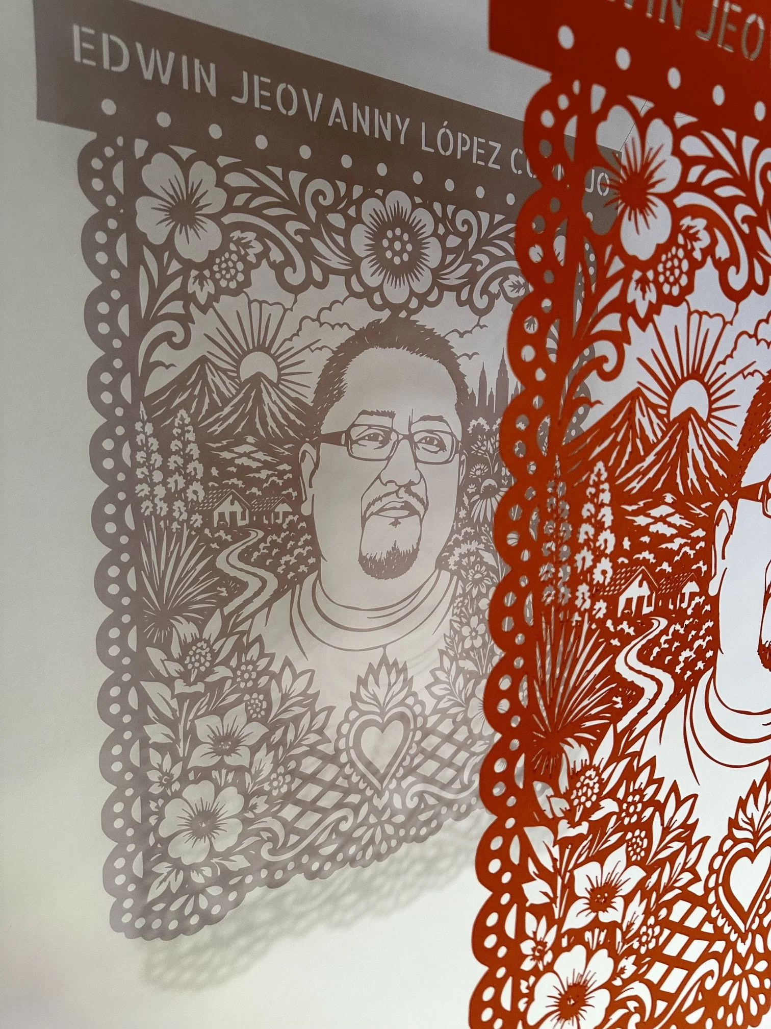 Side view of Edwin Jeovanny López Cornejo’s orange papel picado portrait hanging against a wall, with the cut paper design casting a detailed shadow.
