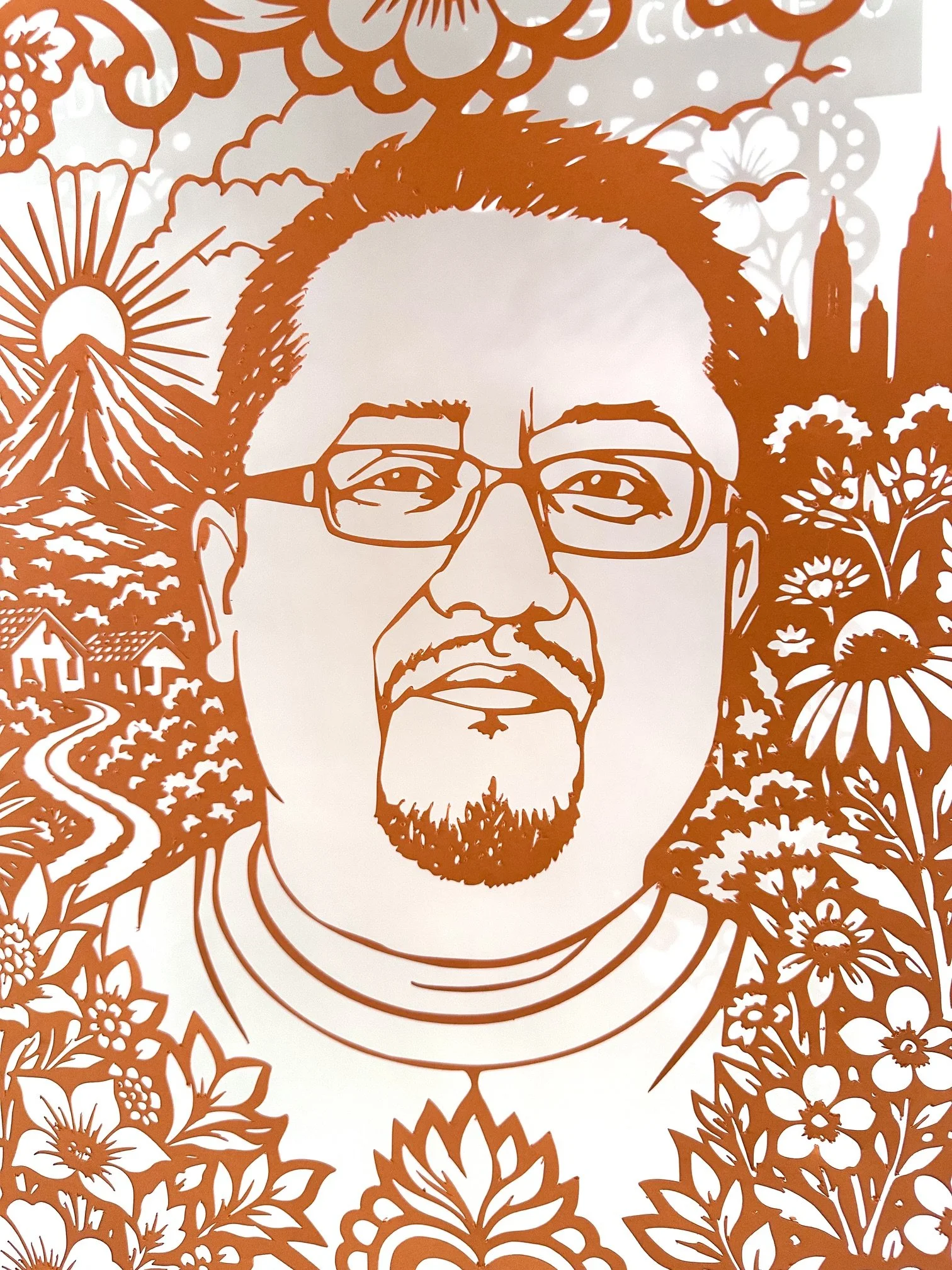 Close up of Edwin Jeovanny López Cornejo’s orange papel picado portrait, showing his face, glasses, mountain landscape, the Manhattan skyline seen from New Jersey and surrounding flowers.