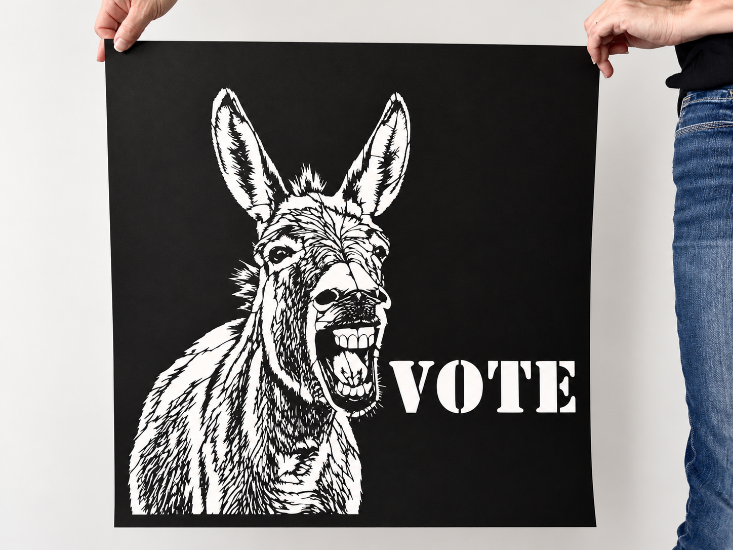 Ruthie political donkey VOTE poster, original black cut paper wall art being held up by two hands showing the scale against a neutral studio wall.