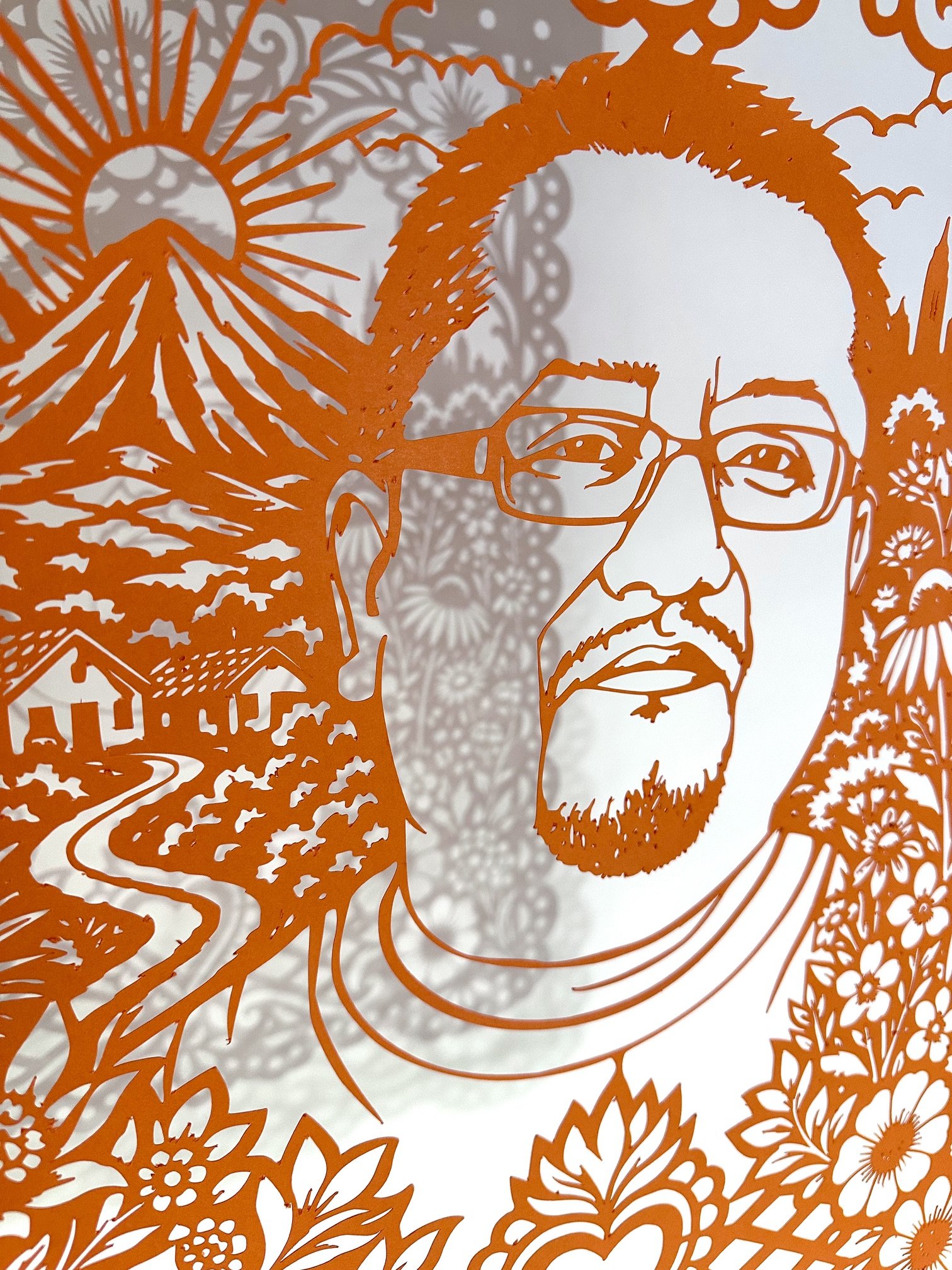 Close view of Edwin Jeovanny López Cornejo’s orange papel picado portrait, showing his face beside a mountain landscape, flowers, and the shadow of the cut paper design.