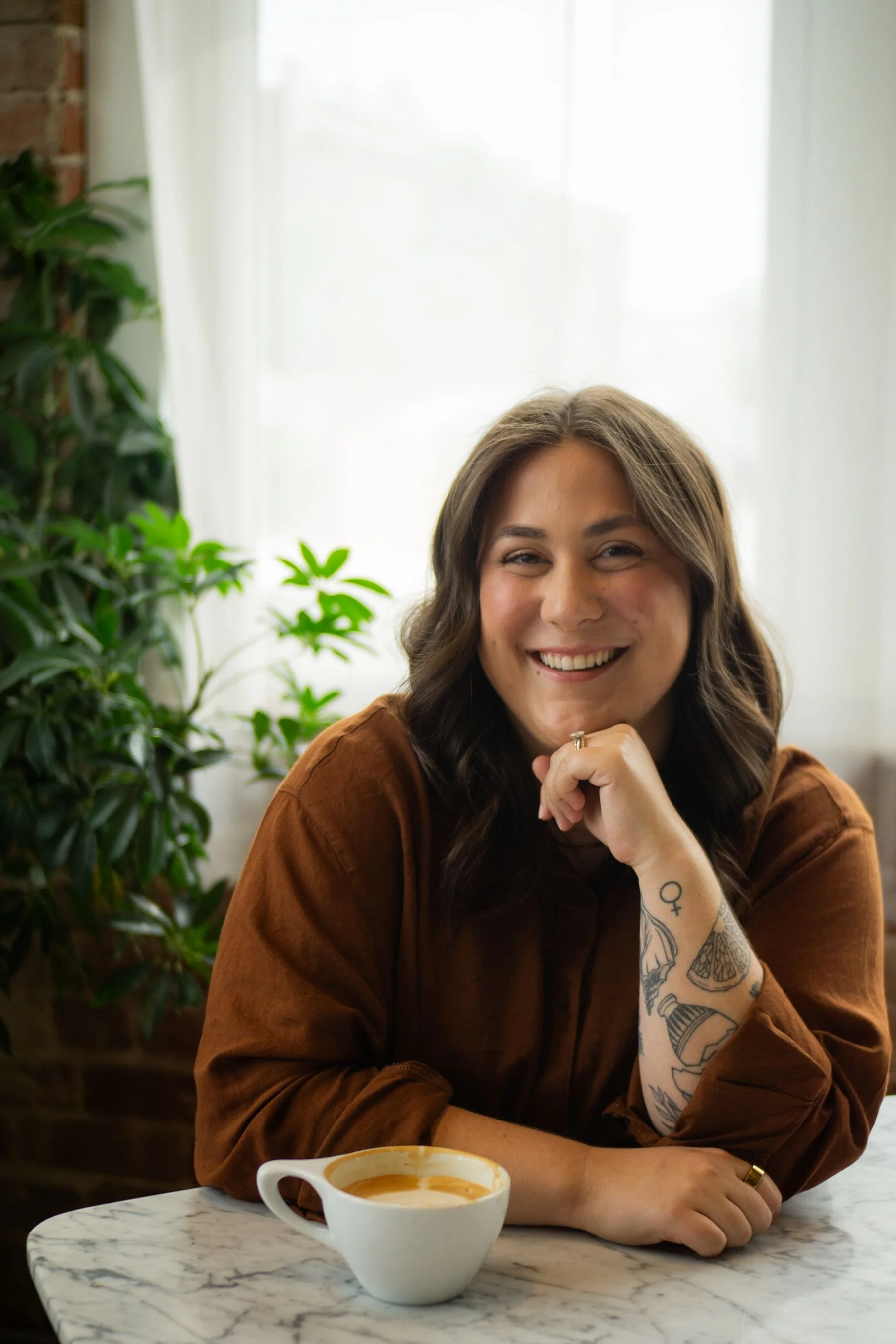 Caroline Kerr, a trauma therapist in Seattle Washington,  with brown hair and tattoos on her arm sitting at a marble table with a cup of coffee, in a cozy cafe with greenery in the background.