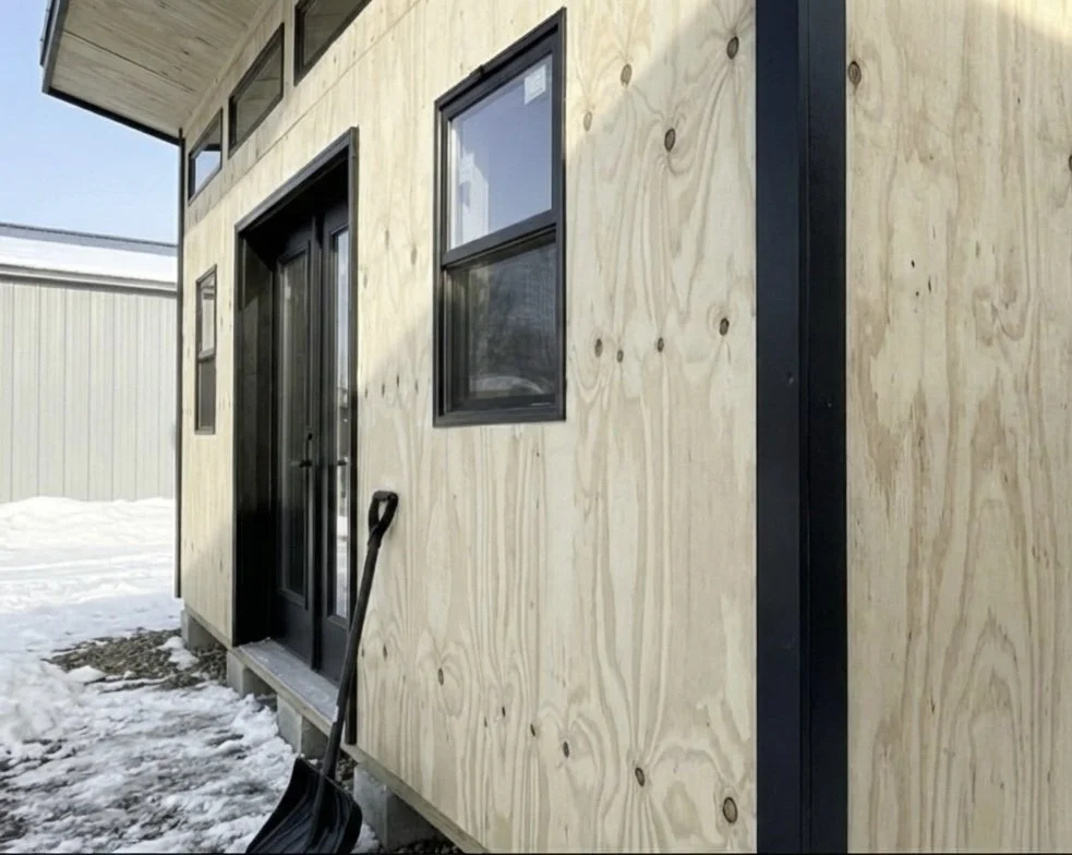 Exterior of a building under construction with plywood siding, black-framed windows, and a black door, with a snow-covered ground and a snow shovel leaning against the wall.