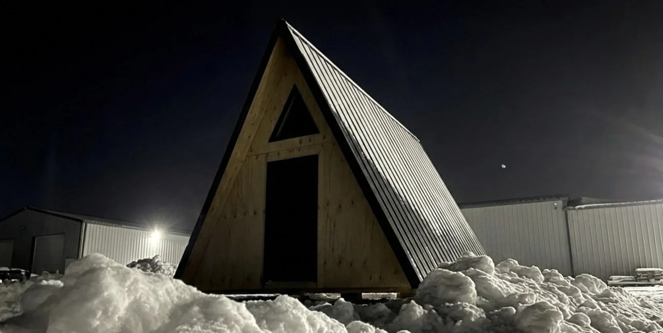 A small wooden A-frame cabin in a snowy setting at night with industrial buildings in the background.