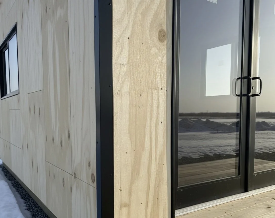 Exterior of a building with plywood siding and glass sliding door, partially covered with snow on the ground.
