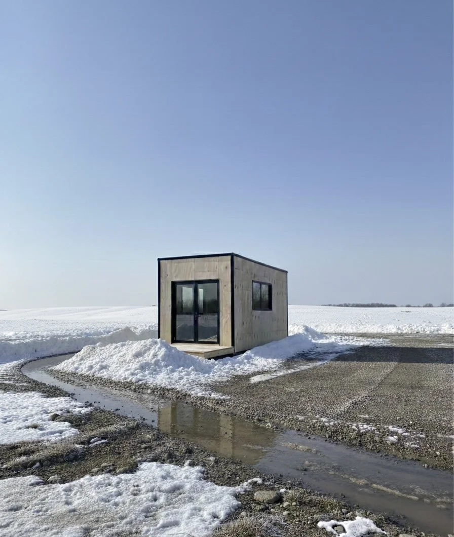 A small wooden structure with glass doors and windows in a snow-covered landscape, next to a wet gravel pathway under a clear sky.