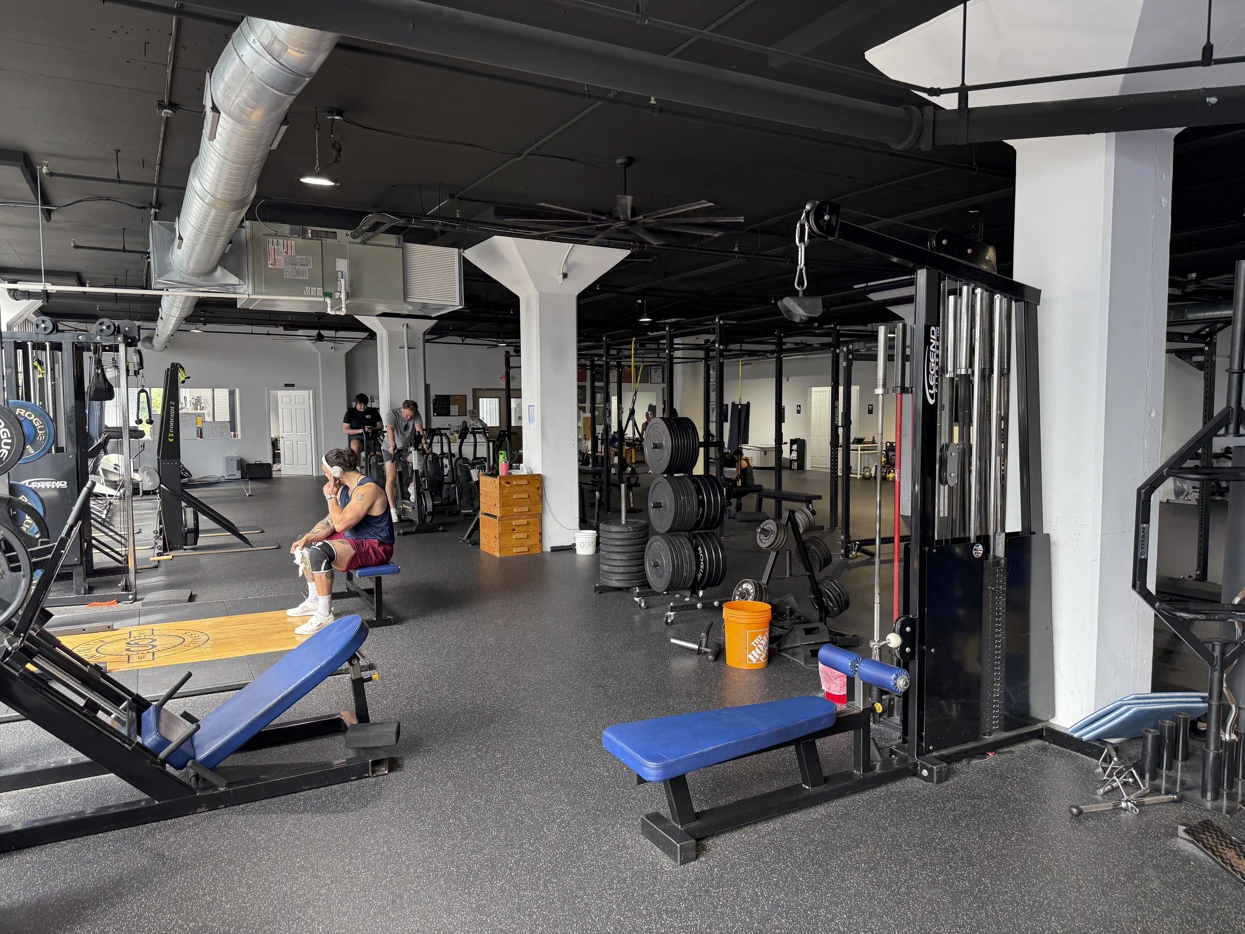 Interior of a gym with various workout equipment, a person sitting on a bench, and others working out in the background.