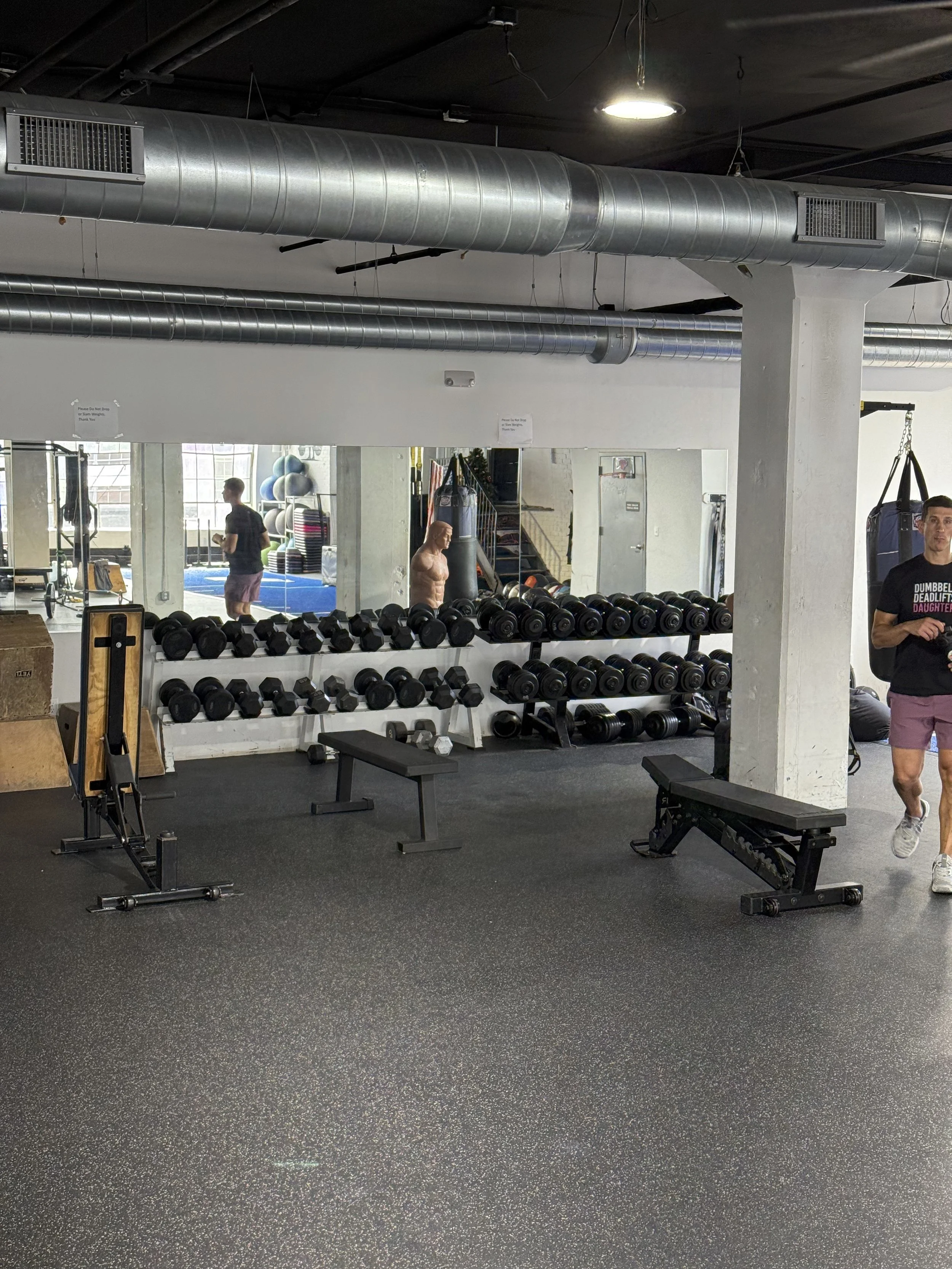 Interior of a gym with weights on racks, benches, and a punching bag, with a person walking and mirrors on the wall.
