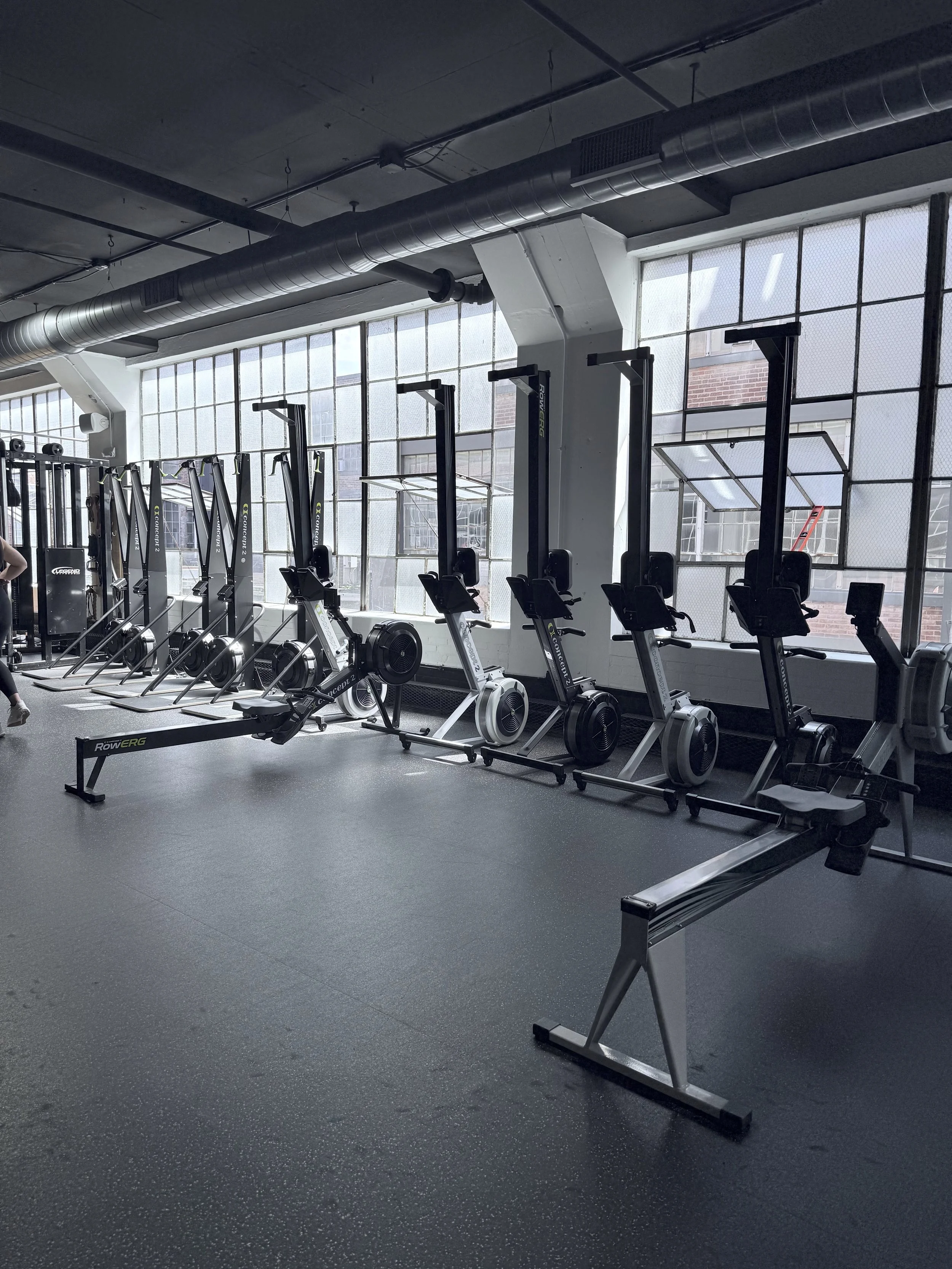 Row of rowing machines in a gym with large windows letting in natural light.