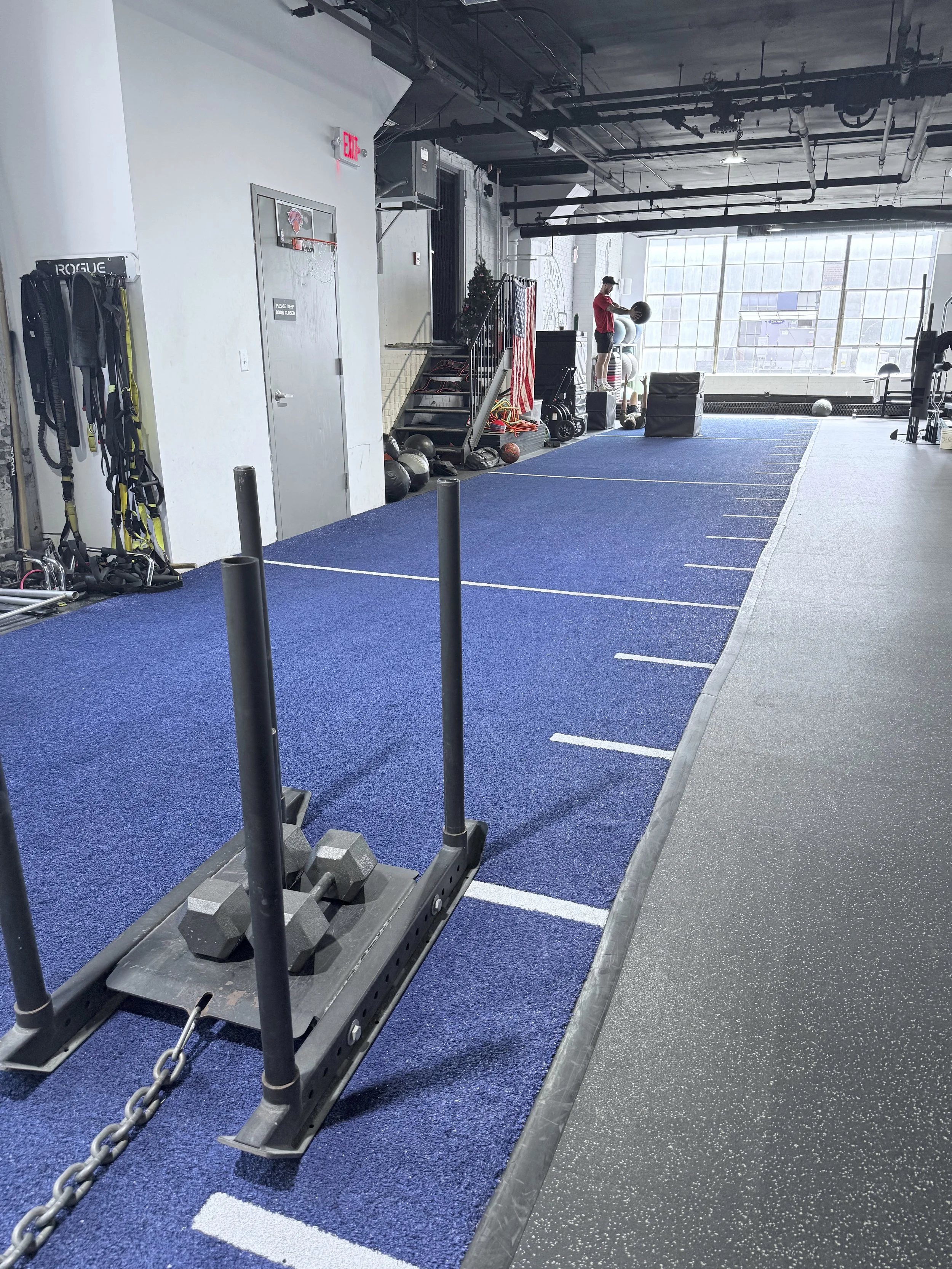 Empty fitness gym with blue turf floor, weight equipment, and a person in a red shirt working out in front of large windows.