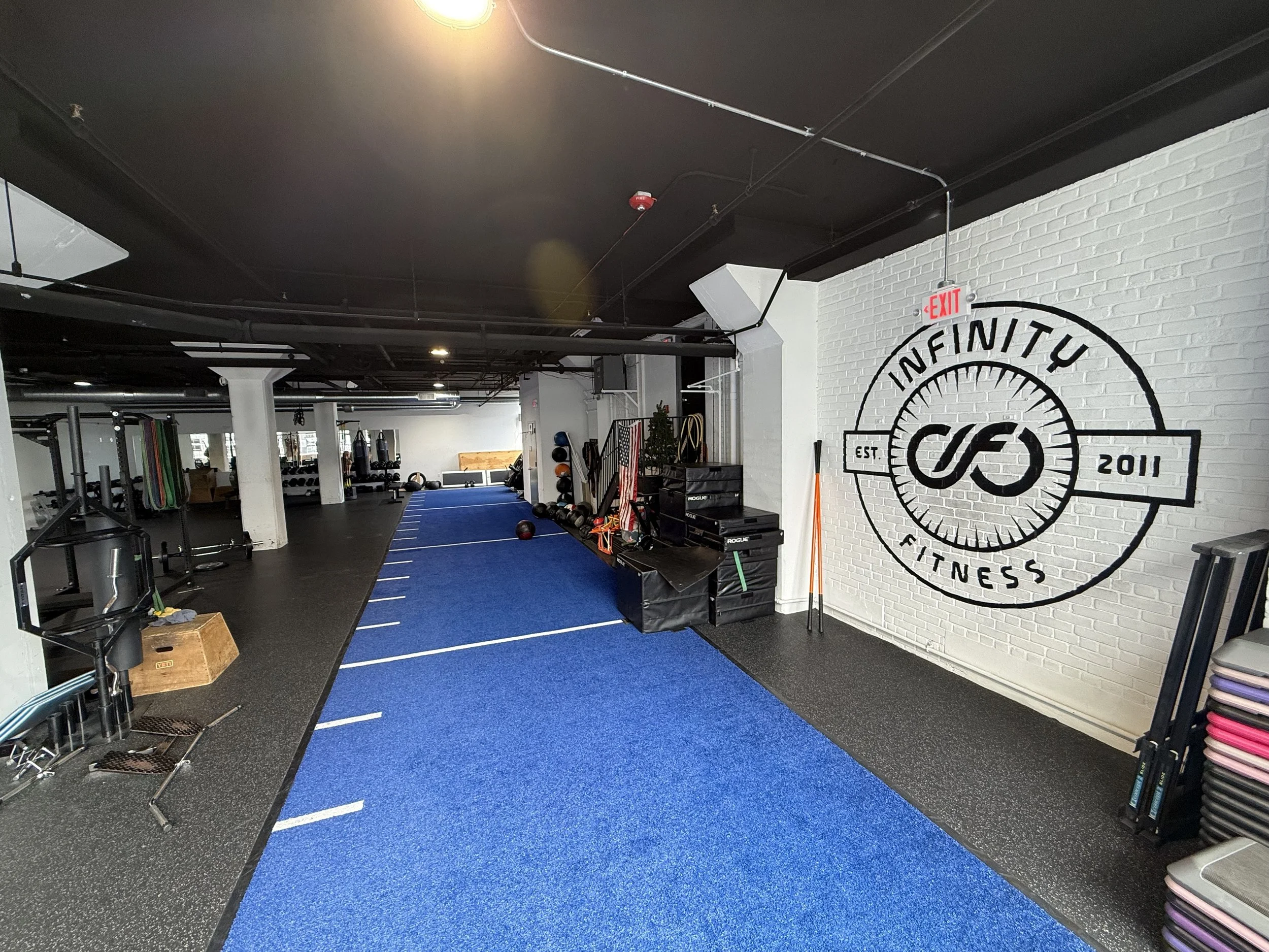 Fitness gym with blue turf area, equipment, and a white brick wall featuring a large logo that reads 'Infinite Fitness' and includes the establishment year 2011.