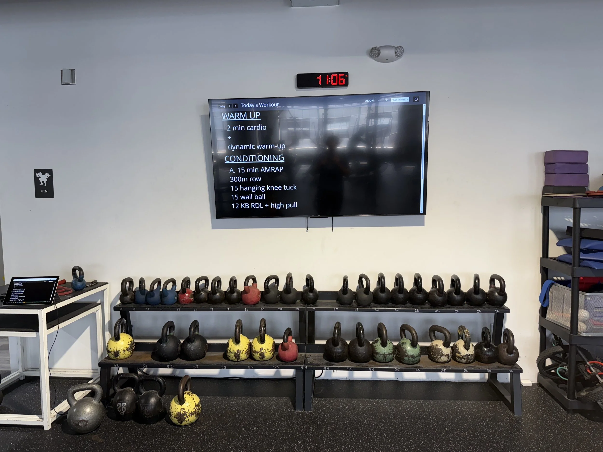 Gym workout area with kettlebells of various colors and sizes arranged on black metal racks, a digital screen displaying workout routines, and a small table with a tablet and fitness accessories.