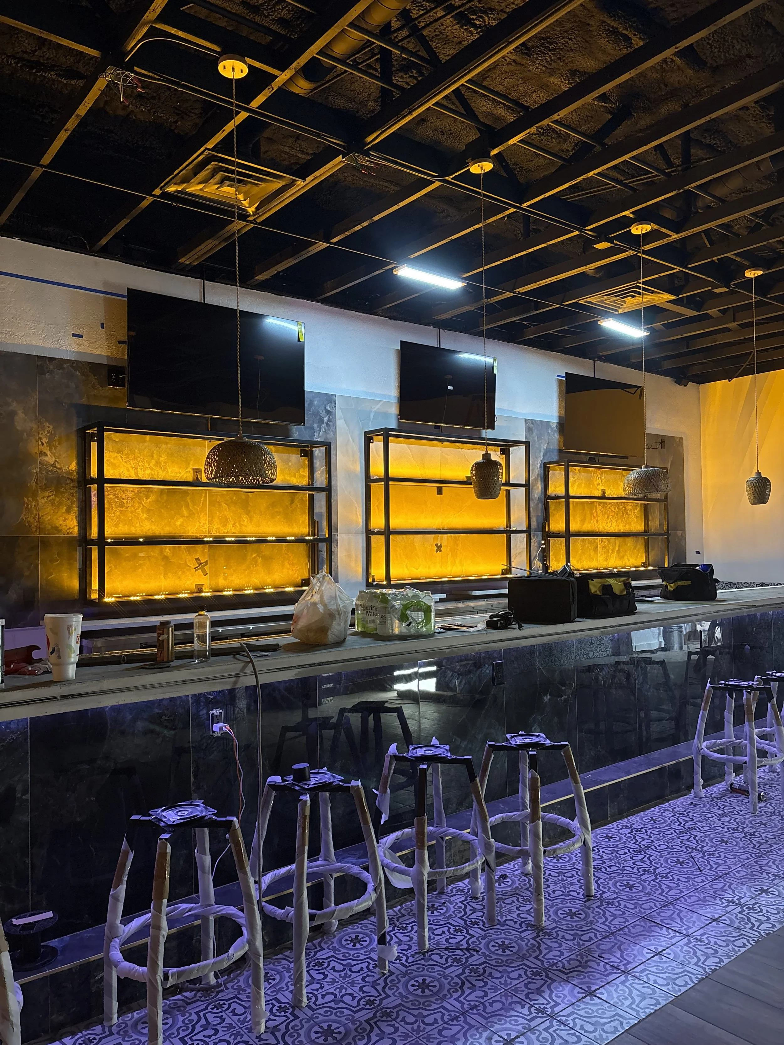Interior of a bar or restaurant under construction with black bar stools, three large wall-mounted TVs, and illuminated shelving units with warm backlighting.