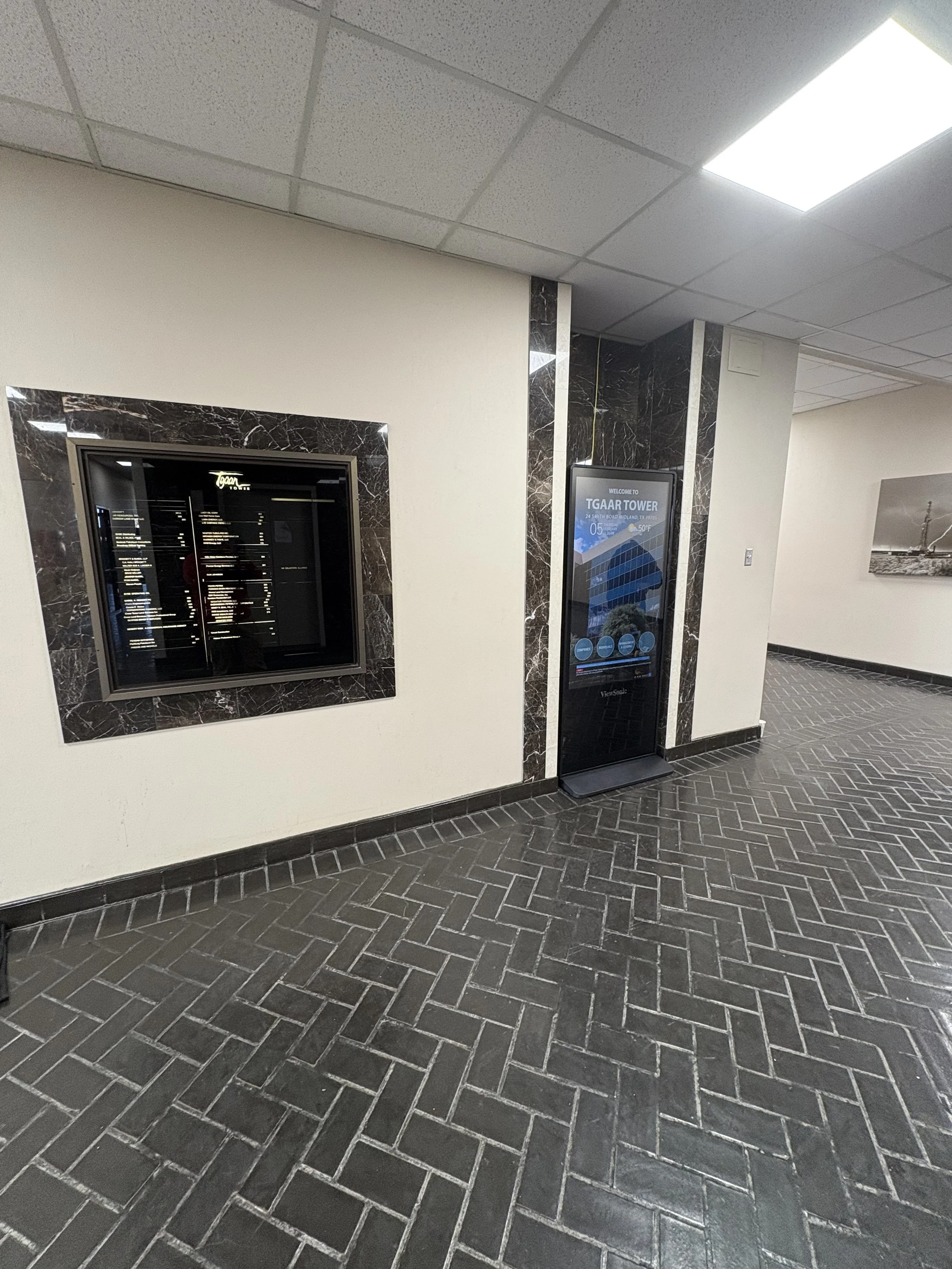 Interior of a building with a wall-mounted directory and an electronic display for TGA Tower, both bordered by black marble trim. The floor features dark herringbone-patterned tile, and there is a painting on the wall in the background.
