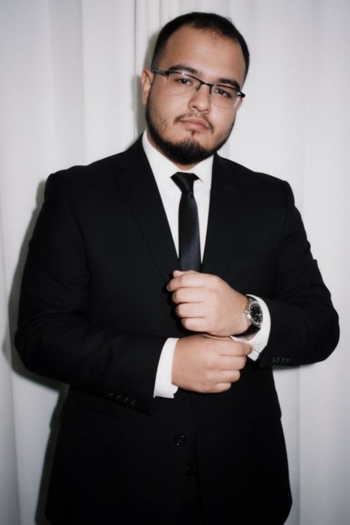 A man in a black suit with a white shirt and black tie, wearing glasses, standing against a white background with his hand adjusting his watch.