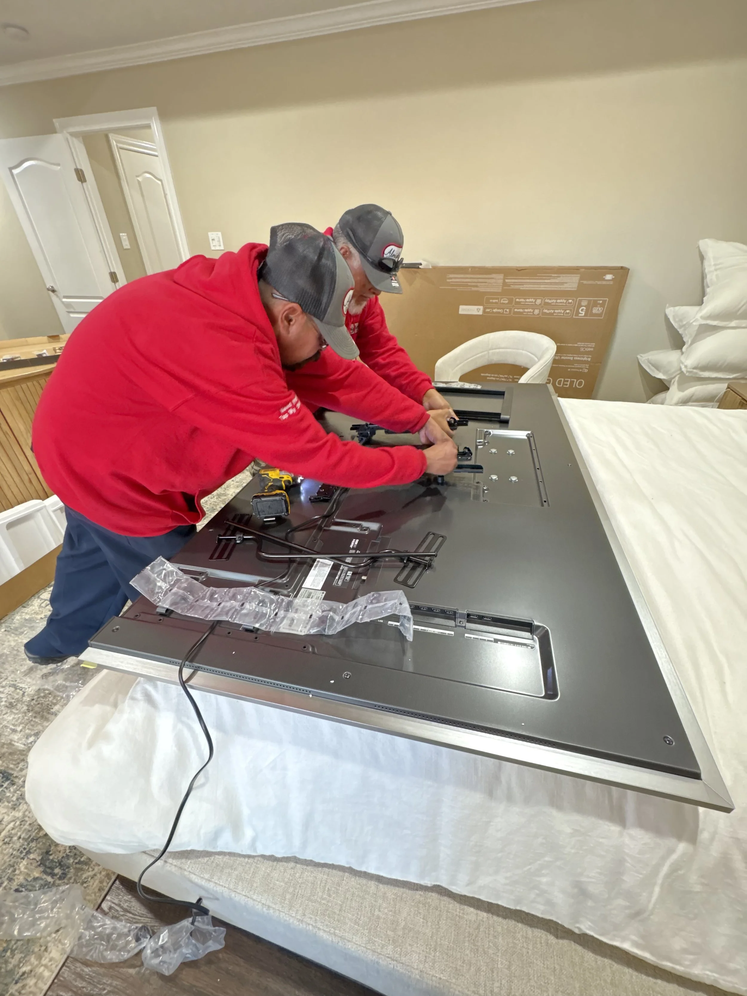 Two men in red hoodies and caps installing or repairing a flat-screen television on a bed in a bedroom.