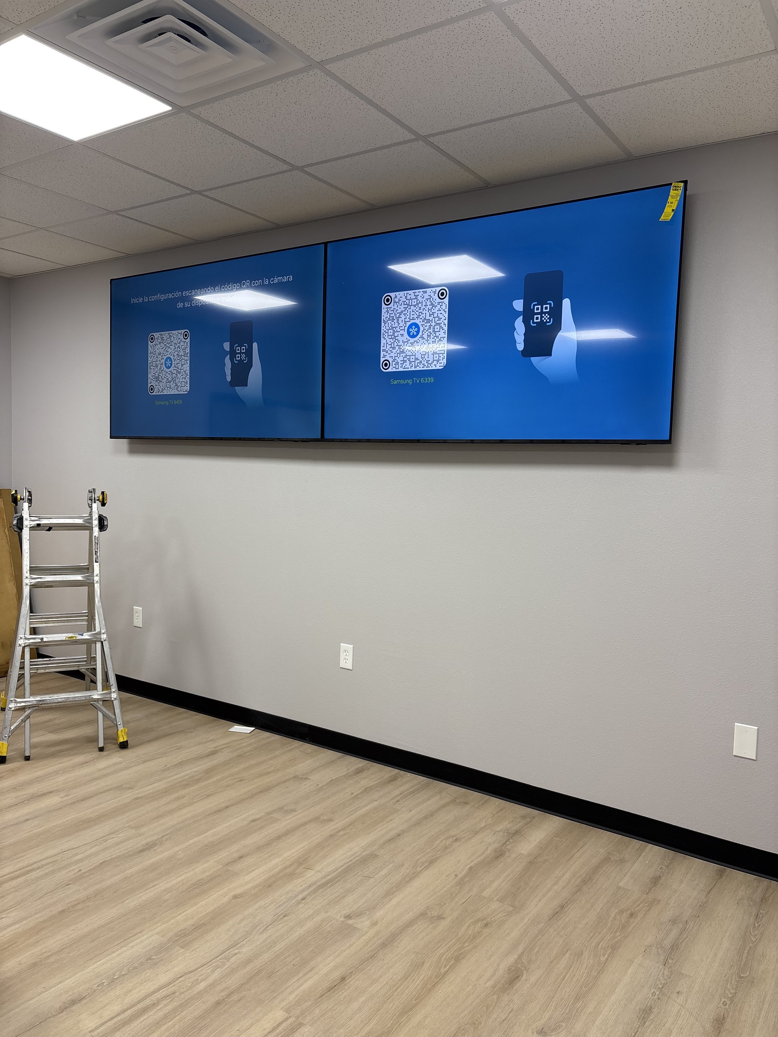 Two large digital screens mounted on a gray wall displaying QR code setup instructions. There is a ladder on the wooden floor in a room with a white ceiling, a light fixture, and electrical outlets.