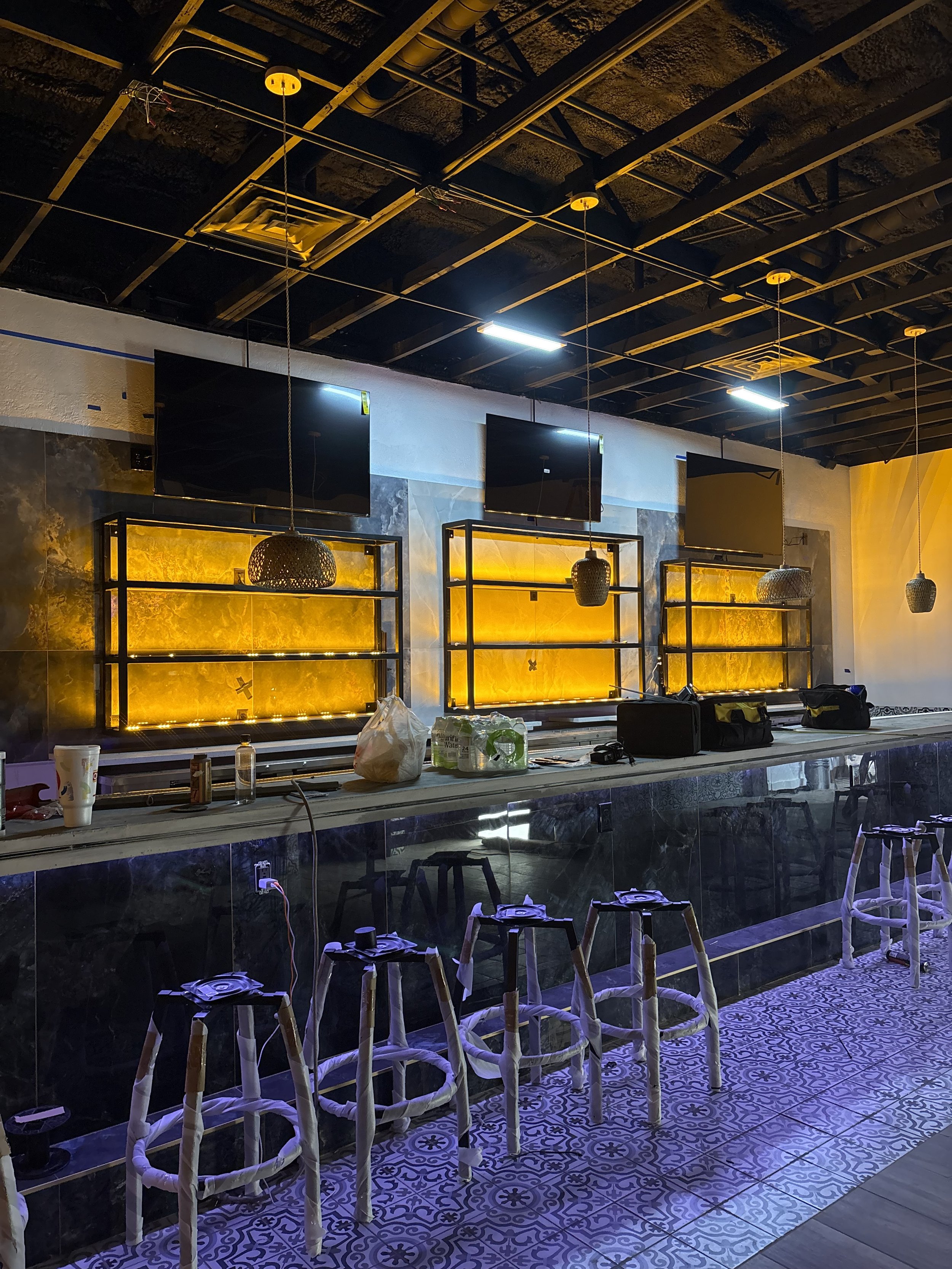 An indoor bar area under construction with barstools lined up at the counter, which has various tools and supplies on it. Behind the counter, there are three large flat screens mounted on the wall, illuminated shelves with yellow backlighting, and hanging pendant lights. The ceiling is exposed with visible ductwork and wiring.