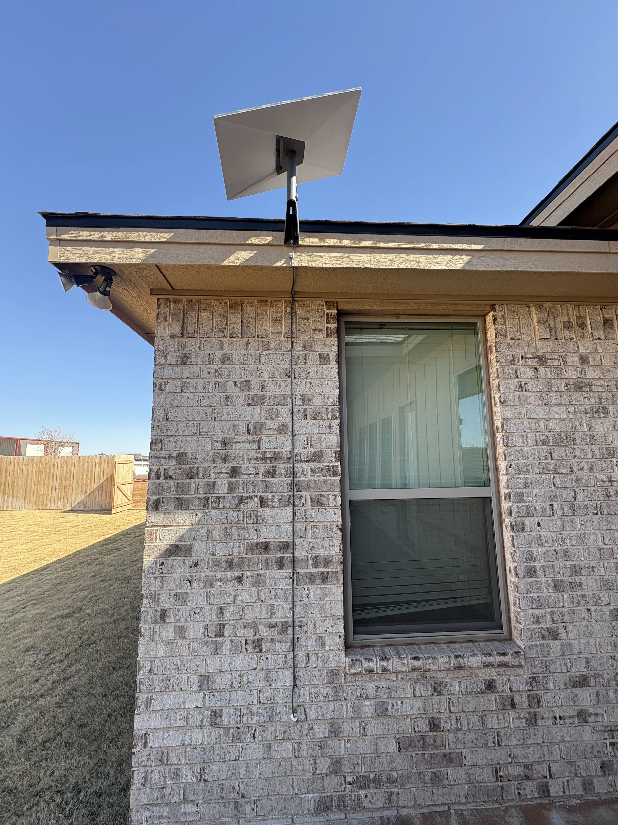 Outside view of a house showing a brick wall, a window with closed blinds, a roof edge, a security camera, and a solar panel mounted on a pole near the roof under a clear blue sky.