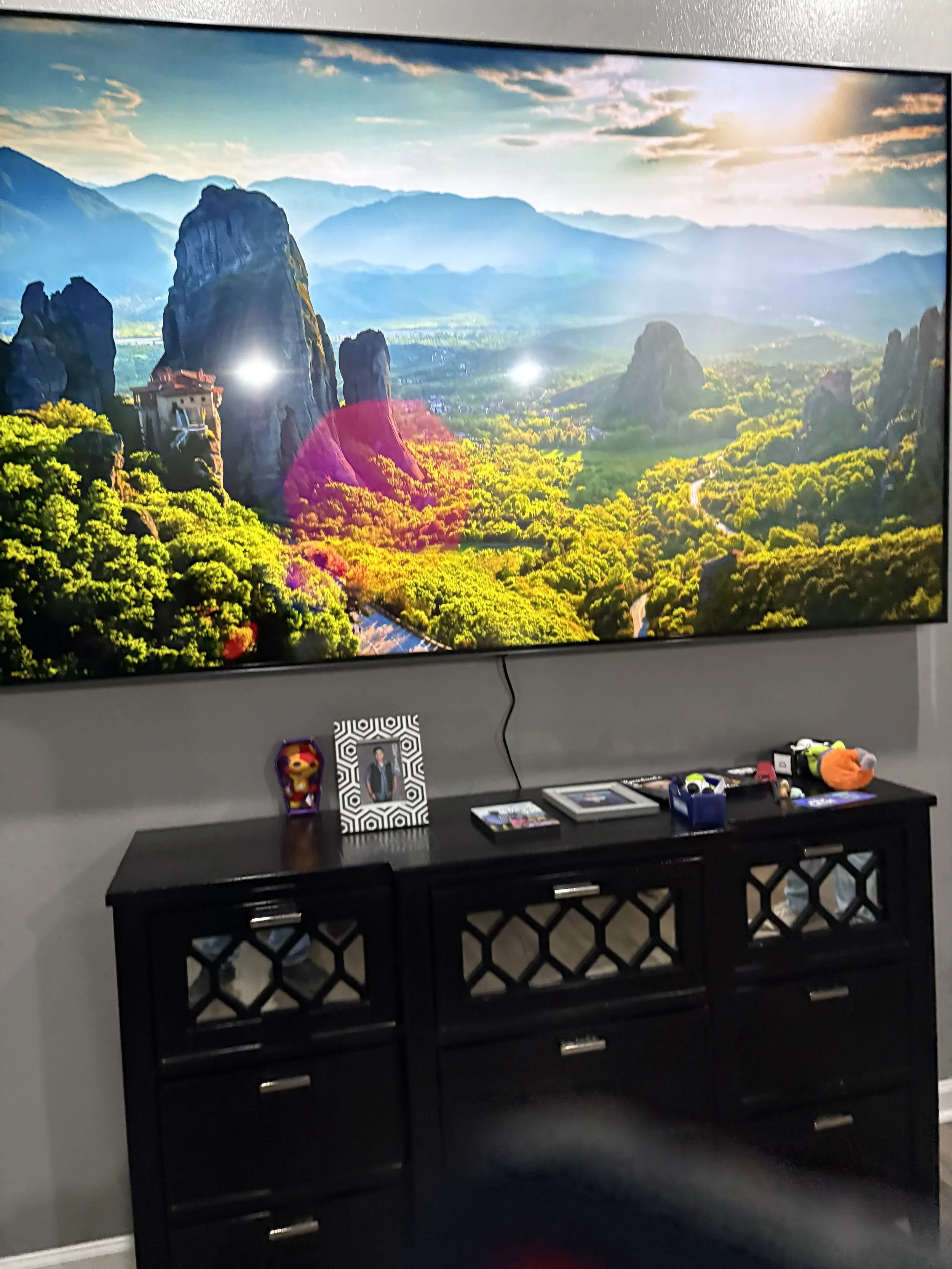 Large flat-screen TV displaying a scenic landscape with mountains, lush green trees, and a sky with clouds and sunlight. In front of the TV is a black console table with family photos, decorative items, and small objects.