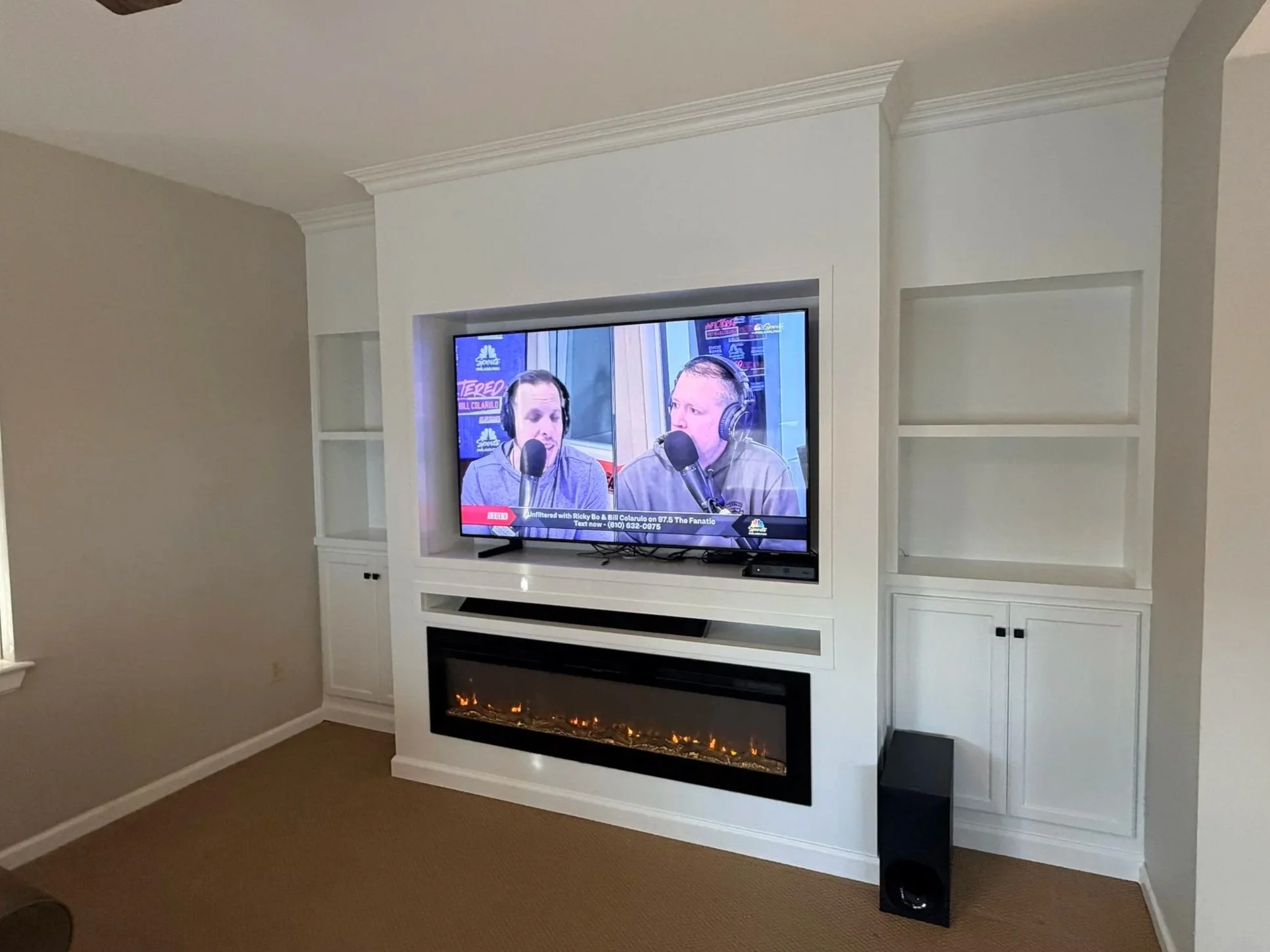 Living room with a wall-mounted flat-screen TV displaying a radio broadcast with two men speaking into microphones, a built-in electric fireplace below the TV, and built-in white shelves on either side of the TV.