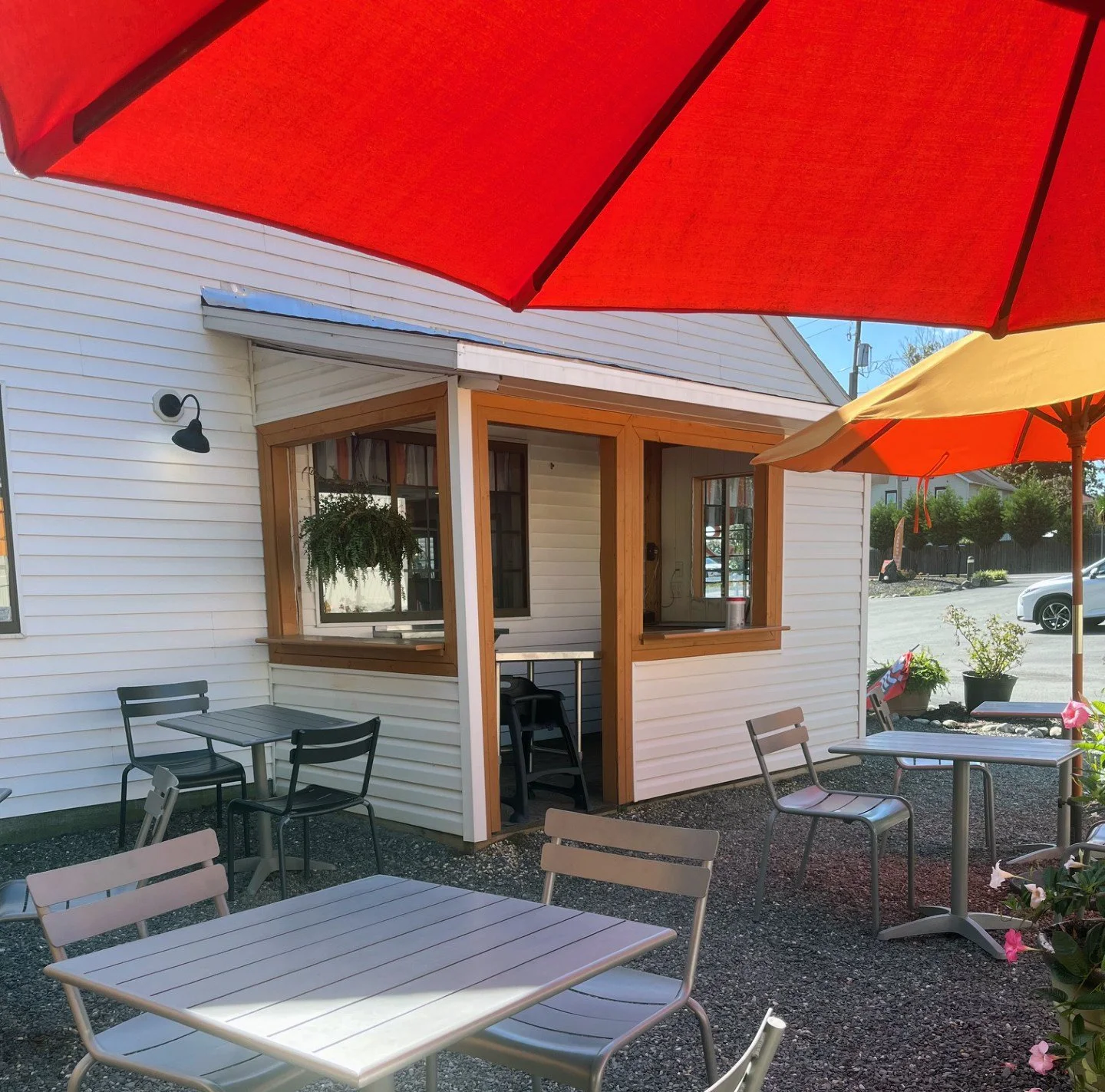 Outdoor patio with tables and chairs under umbrellas outside a white building with wooden window frames and potted plants.
