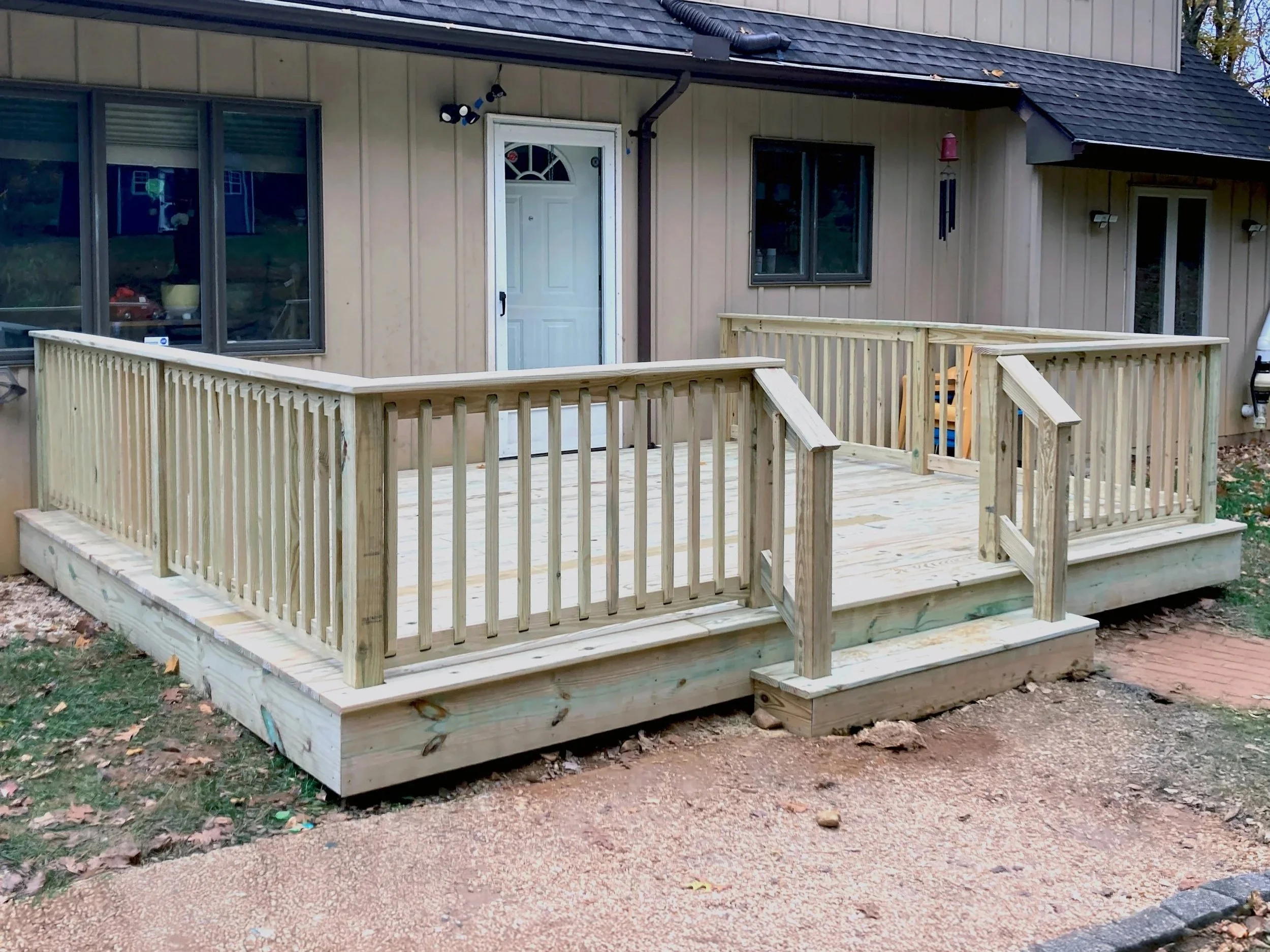 Backyard wooden deck with railings in front of a beige house, with a white door and window, and outdoor lighting fixtures.
