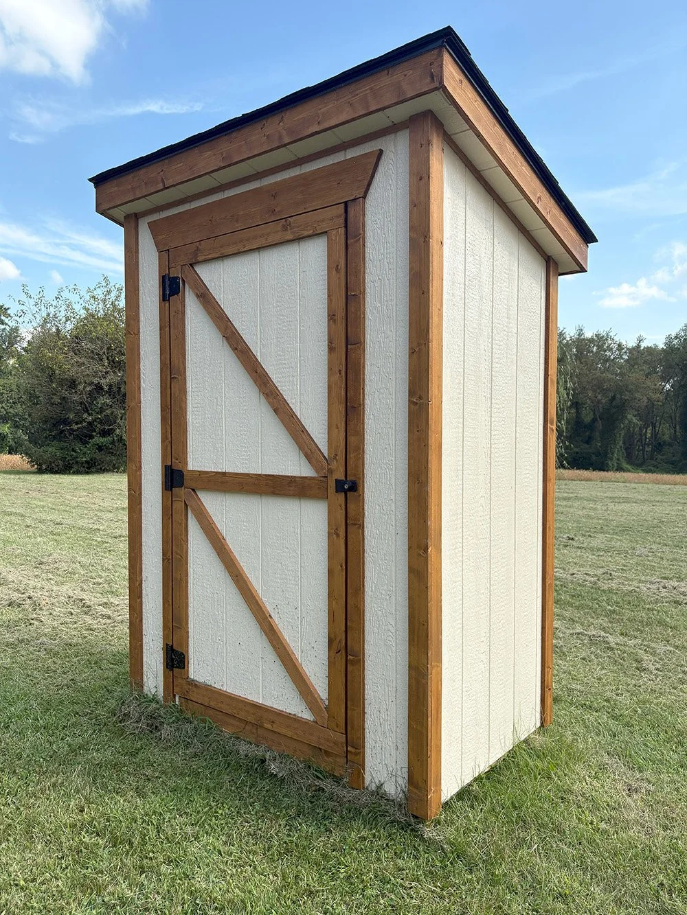 Small shed with white textured walls, wooden trim, and a slanted roof, situated on a grassy field under a blue sky.