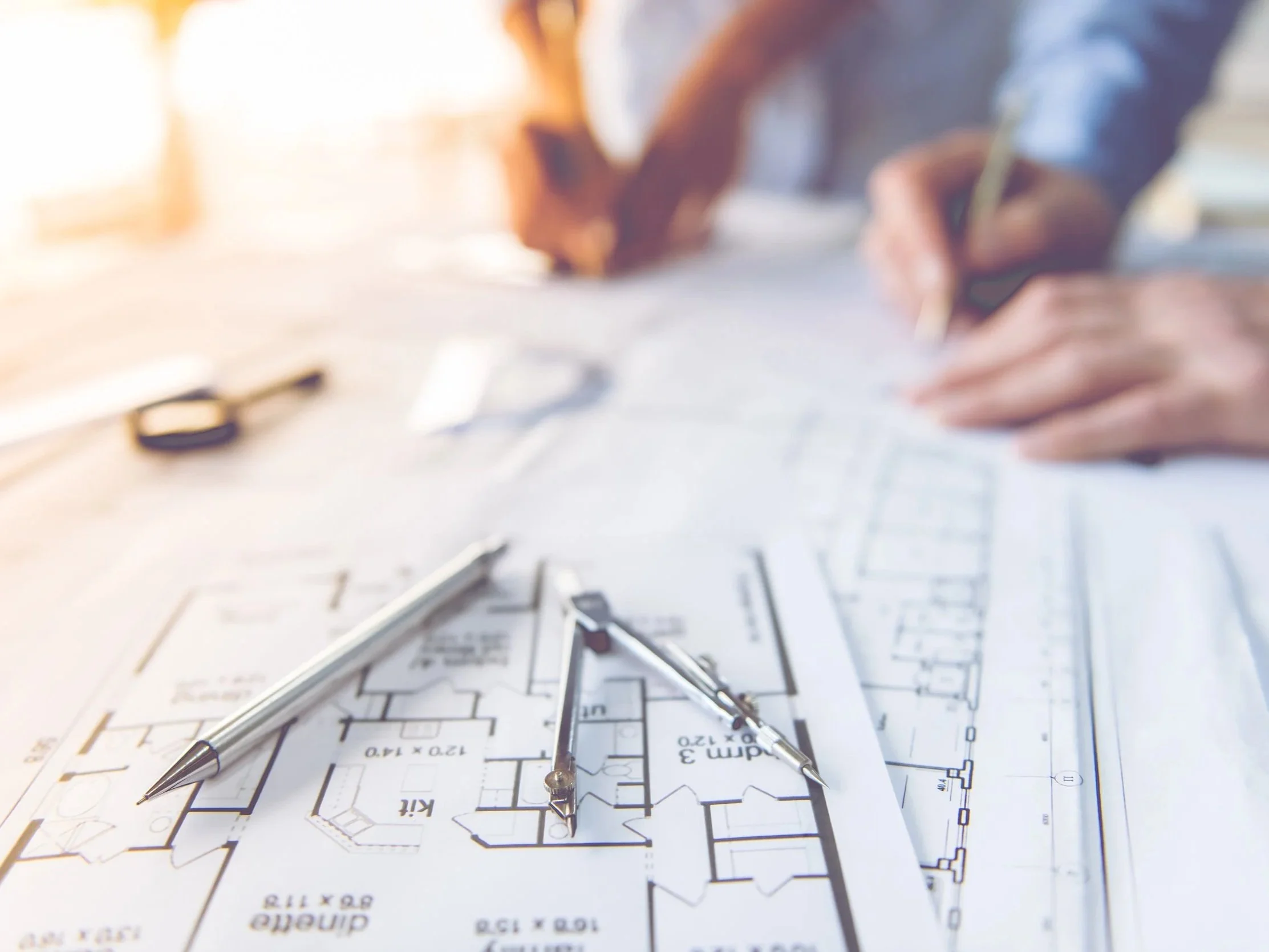 Blueprints and drafting tools on a desk with a person sketching in the background.