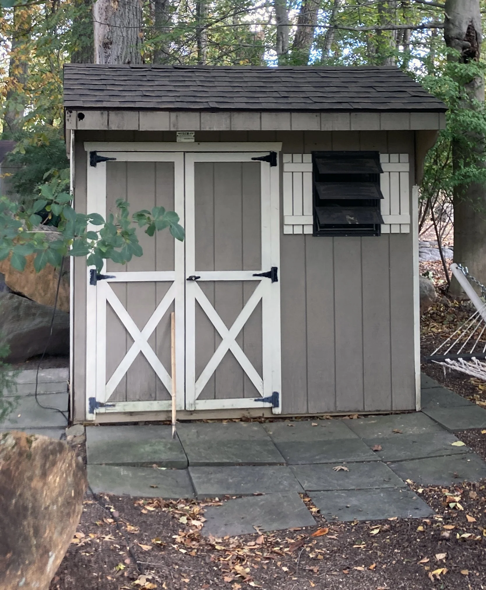 A small gray garden shed with a shingled roof, white double doors with black hinges, a wooden handle, and a small wooden stick leaning against the door. To the right of the shed is a black window with shutters. The shed is surrounded by trees and fol