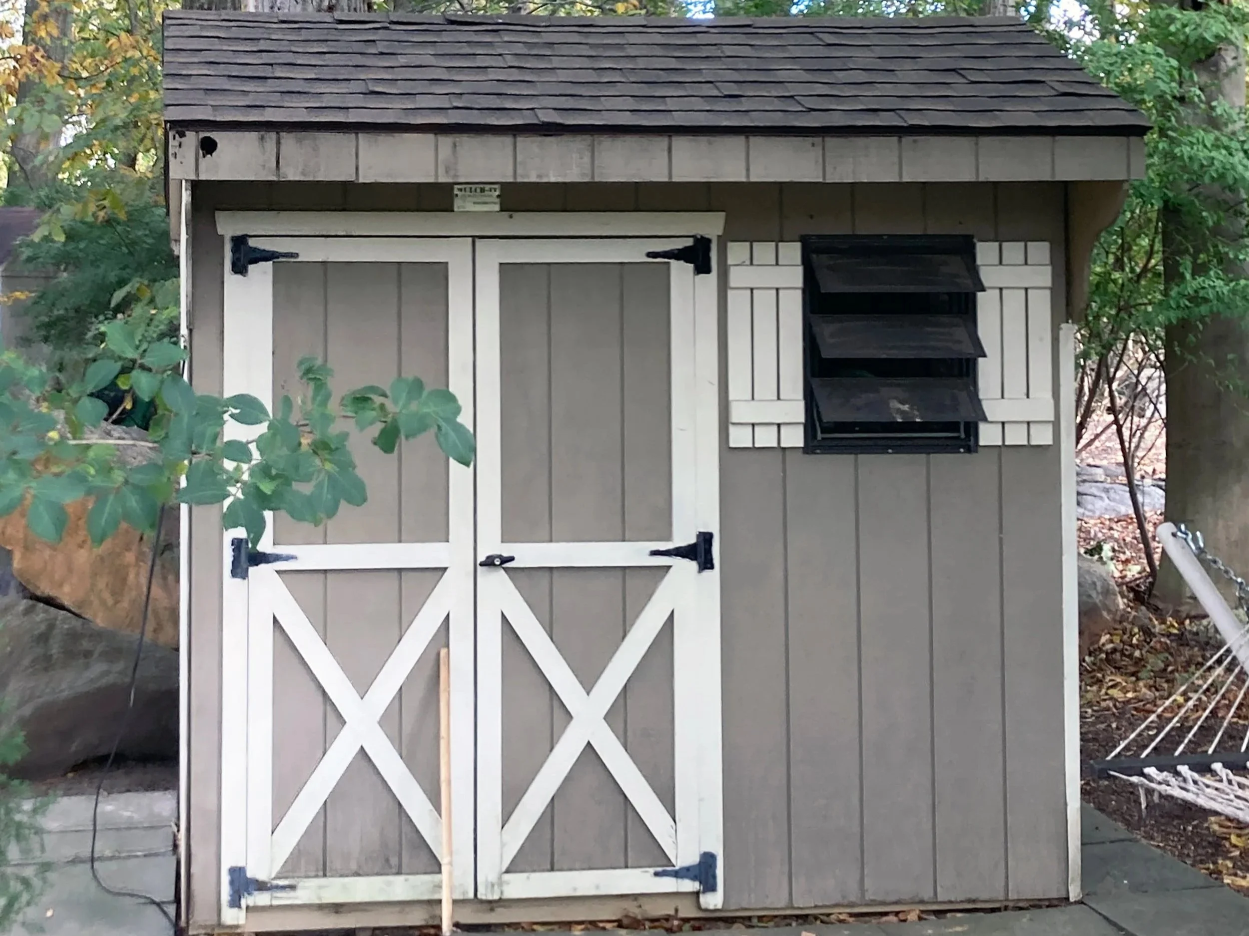 A small outdoor shed with gray siding, white trim, and brown shingle roof. It has a set of double doors with black hinges and a handle, and a small window with black shutters to the right.