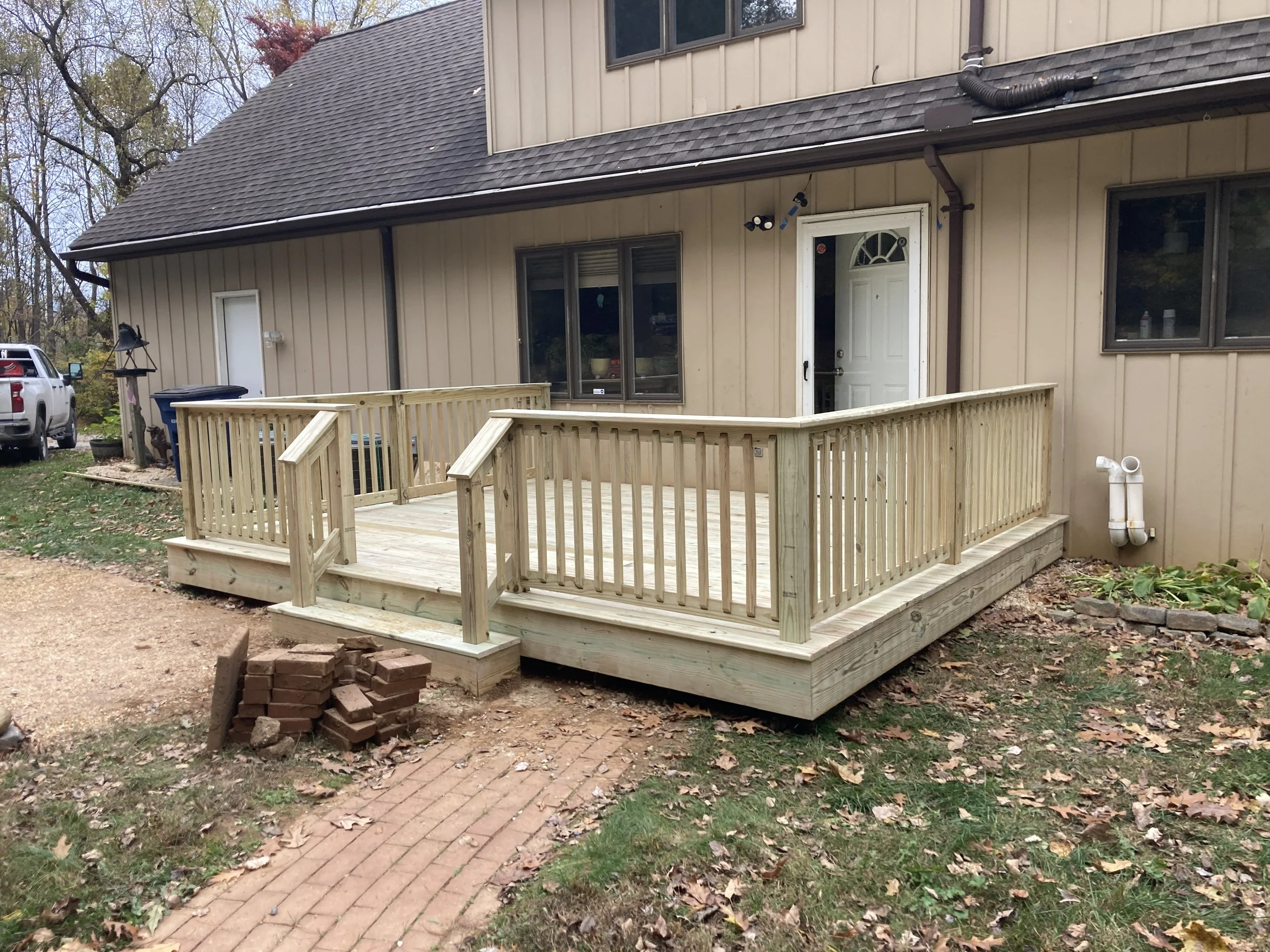 Newly built wooden house deck with stairs and railings in backyard, with landscaping, trees, house exterior, and parked vehicle in background.
