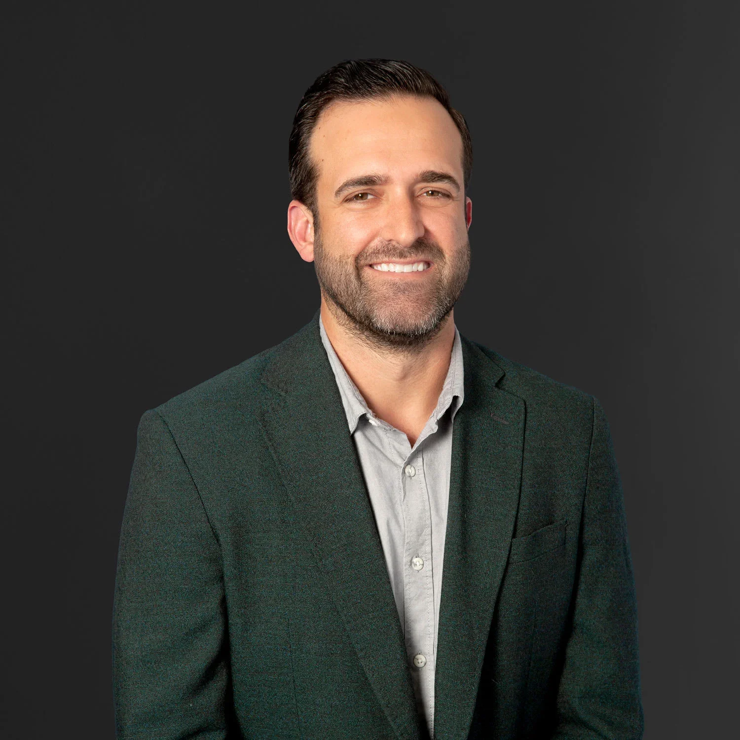 Professional headshot of a man with short dark hair and a beard, wearing a dark blazer and light-colored shirt, smiling against a dark background.