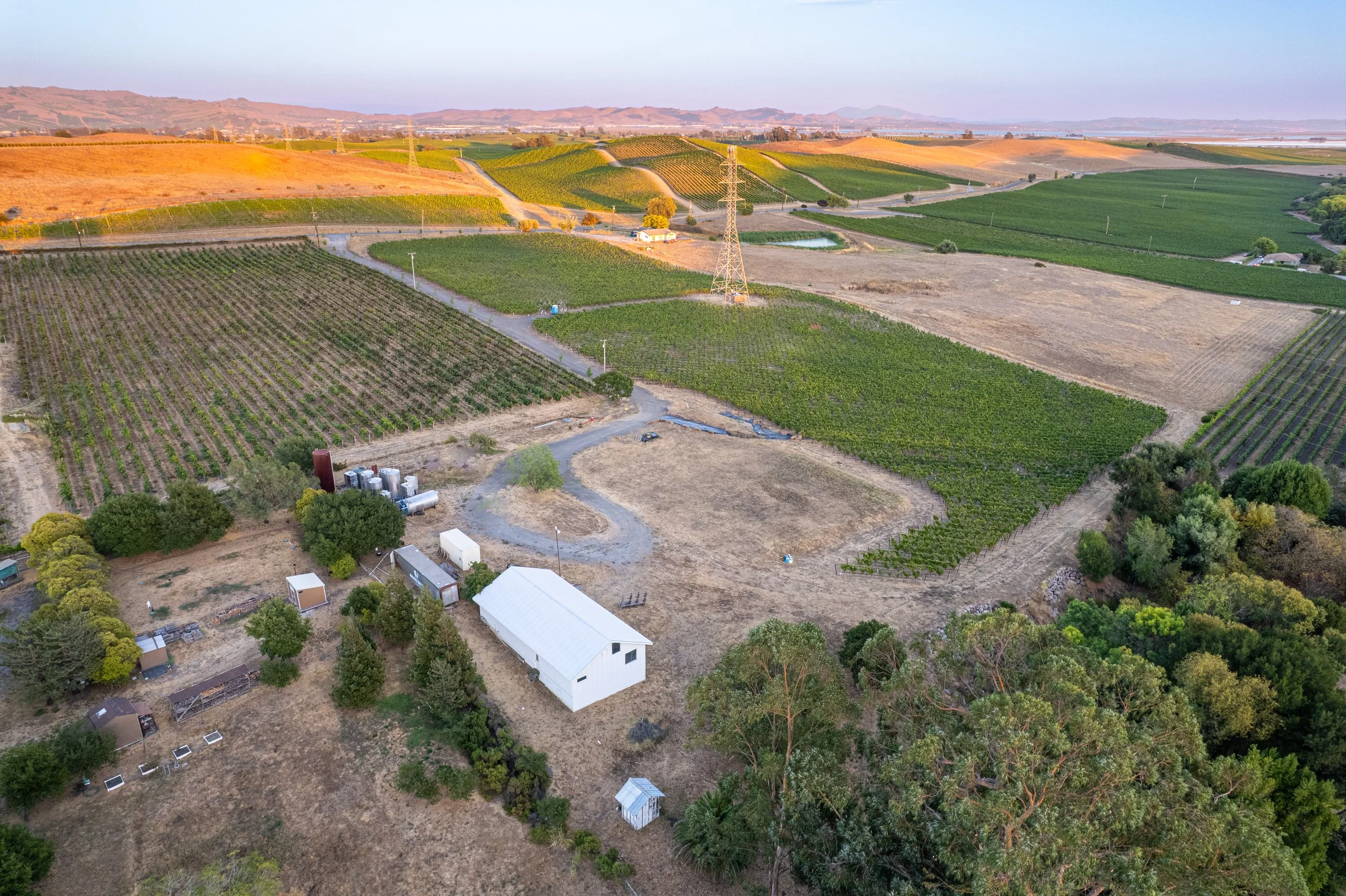 Aerial view of a farm with fields, a white barn, trees, and a dirt road in a rural landscape at sunset.
