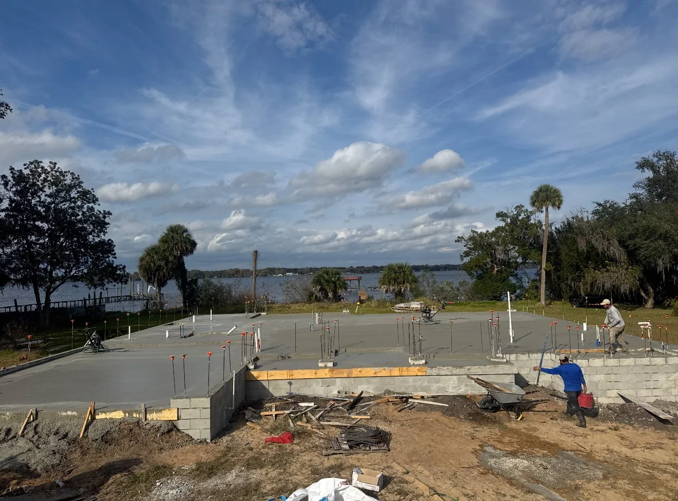 Construction site for a concrete foundation near a body of water with trees and a partly cloudy sky in the background.