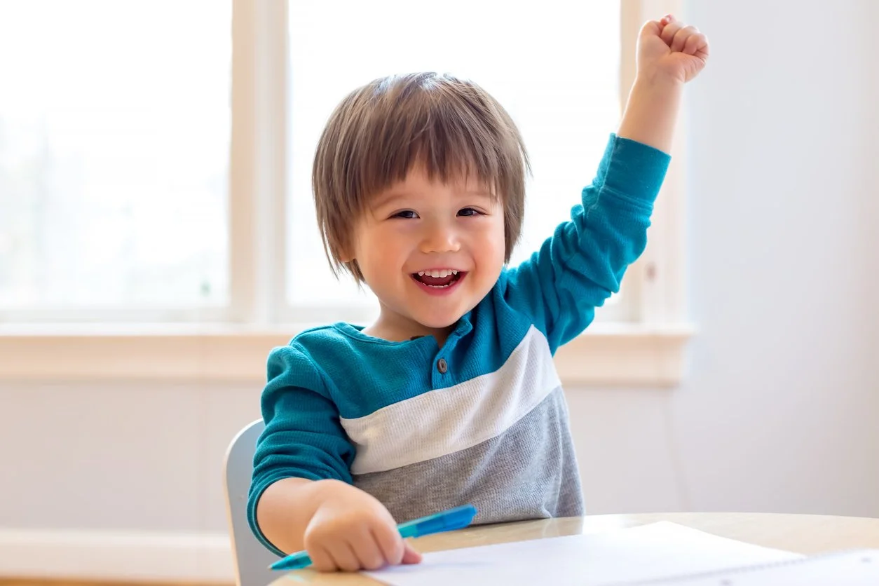 A young boy with brown hair and a bright smile, participating in speech therapy for children in Maui, HI, raising one fist triumphantly while sitting at a table in front of a window with natural light.